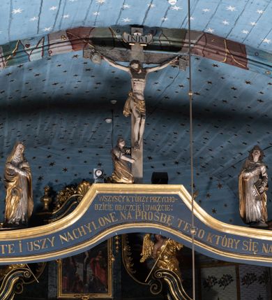 Zdjęcie nr 1: Belka tęczowa o falistym zarysie, zwieńczona profilowanym gzymsem. Z przodu napis „WSZYSCY KTÓRZY PRZYCHO/DZICIE OBACZCIE I UWAŻCIE: / OCZY MOJE BĘDĄ OTWARTE I USZY NACHYLONE NA PROŚBĘ TEGO KTÓRY SIĘ NA TEM MIEJSCU MODLIĆ BĘDZIE.”, na odwrociu napis „TEN KOŚCIÓŁ FUNDOWANY/ PRZEZ BARBARĘ LISICKĄ. / KONSEKROWANY W ROKU 1766. / ODNOMIONY W ROKU 1902. PO RAZ DRUGI W 1980”. Na belce ustawione figury ze sceny Ukrzyżowania. Pośrodku krzyż, do którego przybita jest trzema gwoździami figura umarłego Chrystusa. Ciało w lekkim zwisie o horyzontalnie rozłożonych ramionach, z głową opadającą na prawe ramię, z lekko ugiętymi nogami oraz stopami skrzyżowanymi w układzie prawa na lewą. Twarz ma szczupłą, o zamkniętych oczach, uniesionych brwiach, dużym, wąskim nosie oraz otwartych ustach, okoloną krótką, falowaną brodą i silnie kręconymi włosami opadającymi na ramiona. Na głowie ma koronę cierniową. Ciało jest wychudzone, o silnie zaznaczonych, guzowatych żebrach klatki piersiowej oraz mięśniach i ścięgnach ramion i ud. W partii bioder złocone perizonium zawieszone na sznurze, odsłaniające prawe udo, zawiązane ze zwisem tkaniny na prawym biodrze. 
U dołu krzyża klęczy Maria Magdalena, zwrócona w trzech czwartych w lewo, z uniesioną głową, obiema rękami obejmuje belkę. Twarz ma pociągłą, o małych oczach, krótkim nosie i wąskich ustach, okoloną długimi falowanymi włosami opadającymi na ramiona. Ubrana jest w srebrzoną suknię oraz złocony płaszcz. 
Po prawej stronie krzyża figura Marii, zwrócona w trzech czwartych w lewo, w kontrapoście, z pochyloną głową i rękami skrzyżowanymi na piersi. Twarz ma pociągłą, o wąskich oczach, uniesionych brwiach, małych nosie i ustach. Ubrana jest w srebrzoną suknię oraz złocony płaszcz narzucony na głowę i ramiona, wokół głowy ma białą podwikę. 
Po lewej stronie krzyża św. Jan zwrócony w trzech czwartych w prawo, w kontrapoście, prawą rękę kładzie na piersi, w lewej, opuszczonej trzyma księgę. Twarz ma pociągłą, o wąskich oczach, małych nosie i ustach, okoloną brązowymi, silnie kręconymi włosami, sięgającymi ramion, z czubem nad czołem. Ubrany jest w srebrzoną tunikę z kołnierzykiem, przepasaną w talii oraz złocony płaszcz zarzucony na prawe ramię i podtrzymywany na lewym boku. Polichromia ciał postaci naturalistyczna. 