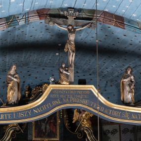 Zdjęcie nr 1: Belka tęczowa o falistym zarysie, zwieńczona profilowanym gzymsem. Z przodu napis „WSZYSCY KTÓRZY PRZYCHO/DZICIE OBACZCIE I UWAŻCIE: / OCZY MOJE BĘDĄ OTWARTE I USZY NACHYLONE NA PROŚBĘ TEGO KTÓRY SIĘ NA TEM MIEJSCU MODLIĆ BĘDZIE.”, na odwrociu napis „TEN KOŚCIÓŁ FUNDOWANY/ PRZEZ BARBARĘ LISICKĄ. / KONSEKROWANY W ROKU 1766. / ODNOMIONY W ROKU 1902. PO RAZ DRUGI W 1980”. Na belce ustawione figury ze sceny Ukrzyżowania. Pośrodku krzyż, do którego przybita jest trzema gwoździami figura umarłego Chrystusa. Ciało w lekkim zwisie o horyzontalnie rozłożonych ramionach, z głową opadającą na prawe ramię, z lekko ugiętymi nogami oraz stopami skrzyżowanymi w układzie prawa na lewą. Twarz ma szczupłą, o zamkniętych oczach, uniesionych brwiach, dużym, wąskim nosie oraz otwartych ustach, okoloną krótką, falowaną brodą i silnie kręconymi włosami opadającymi na ramiona. Na głowie ma koronę cierniową. Ciało jest wychudzone, o silnie zaznaczonych, guzowatych żebrach klatki piersiowej oraz mięśniach i ścięgnach ramion i ud. W partii bioder złocone perizonium zawieszone na sznurze, odsłaniające prawe udo, zawiązane ze zwisem tkaniny na prawym biodrze. 
U dołu krzyża klęczy Maria Magdalena, zwrócona w trzech czwartych w lewo, z uniesioną głową, obiema rękami obejmuje belkę. Twarz ma pociągłą, o małych oczach, krótkim nosie i wąskich ustach, okoloną długimi falowanymi włosami opadającymi na ramiona. Ubrana jest w srebrzoną suknię oraz złocony płaszcz. 
Po prawej stronie krzyża figura Marii, zwrócona w trzech czwartych w lewo, w kontrapoście, z pochyloną głową i rękami skrzyżowanymi na piersi. Twarz ma pociągłą, o wąskich oczach, uniesionych brwiach, małych nosie i ustach. Ubrana jest w srebrzoną suknię oraz złocony płaszcz narzucony na głowę i ramiona, wokół głowy ma białą podwikę. 
Po lewej stronie krzyża św. Jan zwrócony w trzech czwartych w prawo, w kontrapoście, prawą rękę kładzie na piersi, w lewej, opuszczonej trzyma księgę. Twarz ma pociągłą, o wąskich oczach, małych nosie i ustach, okoloną brązowymi, silnie kręconymi włosami, sięgającymi ramion, z czubem nad czołem. Ubrany jest w srebrzoną tunikę z kołnierzykiem, przepasaną w talii oraz złocony płaszcz zarzucony na prawe ramię i podtrzymywany na lewym boku. Polichromia ciał postaci naturalistyczna. 