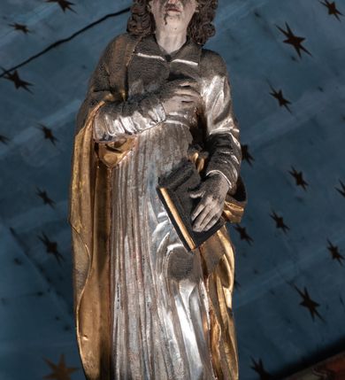 Zdjęcie nr 5: Belka tęczowa o falistym zarysie, zwieńczona profilowanym gzymsem. Z przodu napis „WSZYSCY KTÓRZY PRZYCHO/DZICIE OBACZCIE I UWAŻCIE: / OCZY MOJE BĘDĄ OTWARTE I USZY NACHYLONE NA PROŚBĘ TEGO KTÓRY SIĘ NA TEM MIEJSCU MODLIĆ BĘDZIE.”, na odwrociu napis „TEN KOŚCIÓŁ FUNDOWANY/ PRZEZ BARBARĘ LISICKĄ. / KONSEKROWANY W ROKU 1766. / ODNOMIONY W ROKU 1902. PO RAZ DRUGI W 1980”. Na belce ustawione figury ze sceny Ukrzyżowania. Pośrodku krzyż, do którego przybita jest trzema gwoździami figura umarłego Chrystusa. Ciało w lekkim zwisie o horyzontalnie rozłożonych ramionach, z głową opadającą na prawe ramię, z lekko ugiętymi nogami oraz stopami skrzyżowanymi w układzie prawa na lewą. Twarz ma szczupłą, o zamkniętych oczach, uniesionych brwiach, dużym, wąskim nosie oraz otwartych ustach, okoloną krótką, falowaną brodą i silnie kręconymi włosami opadającymi na ramiona. Na głowie ma koronę cierniową. Ciało jest wychudzone, o silnie zaznaczonych, guzowatych żebrach klatki piersiowej oraz mięśniach i ścięgnach ramion i ud. W partii bioder złocone perizonium zawieszone na sznurze, odsłaniające prawe udo, zawiązane ze zwisem tkaniny na prawym biodrze. 
U dołu krzyża klęczy Maria Magdalena, zwrócona w trzech czwartych w lewo, z uniesioną głową, obiema rękami obejmuje belkę. Twarz ma pociągłą, o małych oczach, krótkim nosie i wąskich ustach, okoloną długimi falowanymi włosami opadającymi na ramiona. Ubrana jest w srebrzoną suknię oraz złocony płaszcz. 
Po prawej stronie krzyża figura Marii, zwrócona w trzech czwartych w lewo, w kontrapoście, z pochyloną głową i rękami skrzyżowanymi na piersi. Twarz ma pociągłą, o wąskich oczach, uniesionych brwiach, małych nosie i ustach. Ubrana jest w srebrzoną suknię oraz złocony płaszcz narzucony na głowę i ramiona, wokół głowy ma białą podwikę. 
Po lewej stronie krzyża św. Jan zwrócony w trzech czwartych w prawo, w kontrapoście, prawą rękę kładzie na piersi, w lewej, opuszczonej trzyma księgę. Twarz ma pociągłą, o wąskich oczach, małych nosie i ustach, okoloną brązowymi, silnie kręconymi włosami, sięgającymi ramion, z czubem nad czołem. Ubrany jest w srebrzoną tunikę z kołnierzykiem, przepasaną w talii oraz złocony płaszcz zarzucony na prawe ramię i podtrzymywany na lewym boku. Polichromia ciał postaci naturalistyczna. 