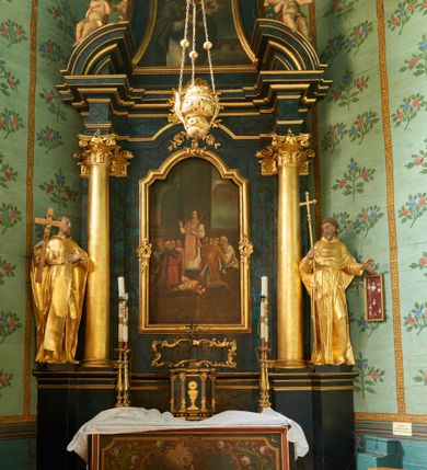 Zdjęcie nr 3: Obraz w formie zbliżonej do trapezu zamkniętego łukiem półkolistym nadwieszonym, o wklęsłych bokach. W centrum św. Jan Nepomucen przedstawiony w półpostaci, zwrócony w trzech czwartych w lewo, z pochyloną głową, z obiema dłońmi złożonymi na piersi, w których trzyma krucyfiks. Twarz ma pociągłą, szczupłą, o dużych, okrągłych oczach skierowanych w dół, wąskim, garbatym nosie i pełnych ustach; okoloną krótką brodą i brązowymi, krótkimi włosami. Na głowie ma czarny biret, a wokół niej nim i wieniec z pięciu gwiazd. Ubrany jest w białą komżę obwiedzioną przy brzegach koronką, almucję zawiązaną pod szyją czerwoną wstążką zakończoną medalionem z wizerunkiem Marii. W tle widoczna scena męczeństwa świętego, gdy zrzucono go z mostu Karola do Wełtawy. Powyżej ukazane nocne, zachmurzone niebo, rozjaśnione na żółto wokół głowy świętego. 
