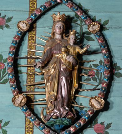 Zdjęcie nr 1: Rzeźba Marii z Dzieciątkiem w mandorli, zwieńczona półpostacią Boga Ojca. Maria ukazana frontalnie, w całej postaci, stojąca na globie i sierpie księżyca, lewą ręką podtrzymuje Dzieciątko, prawą, opuszczoną wyciąga w bok. Twarz ma owalną, o małych oczach, krótkim nosie i pełnych ustach, okoloną długimi, jasnymi włosami, opadającymi na ramiona. Na głowie ma koronę otwartą. Ubrana jest w długą, różową suknię, przepasaną w talii oraz złocony płaszcz, zapięty pod szyją, opadający na ramiona i zawiązany z przodu postaci. Dzieciątko w pozycji siedzącej, zwrócone w trzech czwartych w prawo, z głową skierowaną w przeciwną stronę. Prawą ręką podtrzymuje jabłko królewskie, lewą unosi w geście błogosławieństwa. Twarz ma pociągłą, o małych oczach, krótkim nosie i małych ustach, okoloną kręconymi, jasnymi włosami. Na głowie ma koronę otwartą. Ubrane jest w złoconą tunikę. Figura Marii z Dzieciątkiem w glorii promienistej, ujęta migdałowatą mandorlą, dekorowaną rozetami w kolorach czerwonym i niebieskim, z czterema złoconymi medalionami z przedstawieniami ran Chrystusa – dłoni i stóp. Nad mandorlą półpostać Boga Ojca unoszącego się na obłoku, ukazanego frontalnie. W prawej ręce trzyma jabłko królewskie, lewą unosi w geście błogosławieństwa. Twarz ma podłużną, z małymi oczami, krótkim nosem, okoloną długą brodą oraz długimi, siwymi, kręconymi włosami. Ubrany jest w srebrzoną tunikę, przepasaną w talii oraz złocony płaszcz zapięty pod szyją i opadający na ramiona. 