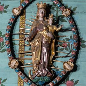 Zdjęcie nr 1: Rzeźba Marii z Dzieciątkiem w mandorli, zwieńczona półpostacią Boga Ojca. Maria ukazana frontalnie, w całej postaci, stojąca na globie i sierpie księżyca, lewą ręką podtrzymuje Dzieciątko, prawą, opuszczoną wyciąga w bok. Twarz ma owalną, o małych oczach, krótkim nosie i pełnych ustach, okoloną długimi, jasnymi włosami, opadającymi na ramiona. Na głowie ma koronę otwartą. Ubrana jest w długą, różową suknię, przepasaną w talii oraz złocony płaszcz, zapięty pod szyją, opadający na ramiona i zawiązany z przodu postaci. Dzieciątko w pozycji siedzącej, zwrócone w trzech czwartych w prawo, z głową skierowaną w przeciwną stronę. Prawą ręką podtrzymuje jabłko królewskie, lewą unosi w geście błogosławieństwa. Twarz ma pociągłą, o małych oczach, krótkim nosie i małych ustach, okoloną kręconymi, jasnymi włosami. Na głowie ma koronę otwartą. Ubrane jest w złoconą tunikę. Figura Marii z Dzieciątkiem w glorii promienistej, ujęta migdałowatą mandorlą, dekorowaną rozetami w kolorach czerwonym i niebieskim, z czterema złoconymi medalionami z przedstawieniami ran Chrystusa – dłoni i stóp. Nad mandorlą półpostać Boga Ojca unoszącego się na obłoku, ukazanego frontalnie. W prawej ręce trzyma jabłko królewskie, lewą unosi w geście błogosławieństwa. Twarz ma podłużną, z małymi oczami, krótkim nosem, okoloną długą brodą oraz długimi, siwymi, kręconymi włosami. Ubrany jest w srebrzoną tunikę, przepasaną w talii oraz złocony płaszcz zapięty pod szyją i opadający na ramiona. 