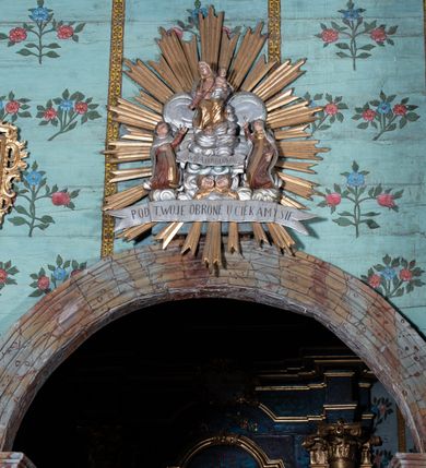 Zdjęcie nr 2: Na tle srebrzonych obłoków przedstawienie tronującej Matki Boskiej z Dzieciątkiem, flankowanej przez klęczące postacie świętych. Maria ukazana jako siedząca, frontalnie, przechylona w prawo, z prawą ręką wyciągniętą w dół oraz lewą podtrzymującą Dzieciątko. Ubrana jest w różową suknię, przepasaną w talii oraz złocony welon i złocony płaszcz, opadający na kolana. Dzieciątko w pozycji siedzącej, zwrócone w lewo, pochylone, obie ręce ma ugięte. Przewiązane jest srebrzoną pieluszką. Po prawej stronie Marii klęczy św. Szymon Stock, zwrócony w trzech czwartych w lewo, z uniesioną głową, odchylony w tył, prawą rękę kładzie na piersi, lewą wyciąga w górę. Twarz ma pełną, na głowie ma tonsurę. Ubrany jest w brązowy habit, ze złoconym szkaplerzem i srebrzonym płaszczem z kapturem. Po lewej stronie klęczy św. Maria Magdalena de Pazzi, zwrócona w trzech czwartych w prawo, z uniesioną głową, lewą rękę ma opuszczoną wzdłuż boku, prawą wyciąga w górę. Ubrana jest w brązowy habit, ze złoconym szkaplerzem oraz w srebrzony welon i płaszcz, na głowie ma zieloną koronę cierniową. U stóp Matki Boskiej, na tle obłoków banderola z napisem „WITAJ KRÓLOWA”, pod nim zaś dwie uskrzydlone główki anielskie. Poniżej całej sceny srebrzona banderola z napisem „POD TWOJA OBRONE UCIEKAMY SIE”. Całość w złoconej glorii promienistej. Polichromia ciał postaci naturalistyczna. 