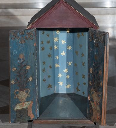 Zdjęcie nr 7: Tabernakulum w formie drewnianej skrzynki, zamkniętej trójbocznie, imitującej budynek, przykrytej wielospadowym dachem, na profilowanym cokole. Drzwiczki dwuskrzydłowe, w kształcie stojącego prostokąta, zwieńczone trójkątnym szczytem, zamocowane na zawiasach z okuciami. Dekorowane malowanymi płycinami o fantazyjnym kształcie i ornamentem.  Ścianki boczne z malowaną architekturą w kolorze jasnobrązowym, dwuosiowe, wydzielone pilastrami dźwigającymi belkowanie. Na osiach malowane okienka w dekoracyjnych opaskach. Na tylnej ściance namalowane belkowanie oraz dwie połówki okien w dekoracyjnych opaskach. Wnętrze tabernakulum polichromowane w kolorze jasnoniebieskim ze złoconymi gwiazdami. Drzwiczki po wewnętrznej stronie z malowaną dekoracją w postaci cokołów z wazonami i kwiatami, na jasnoniebieskim tle, w górnej partii ornament. 
