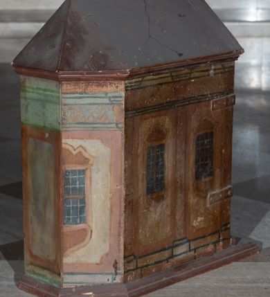Zdjęcie nr 4: Tabernakulum w formie drewnianej skrzynki, zamkniętej trójbocznie, imitującej budynek, przykrytej wielospadowym dachem, na profilowanym cokole. Drzwiczki dwuskrzydłowe, w kształcie stojącego prostokąta, zwieńczone trójkątnym szczytem, zamocowane na zawiasach z okuciami. Dekorowane malowanymi płycinami o fantazyjnym kształcie i ornamentem.  Ścianki boczne z malowaną architekturą w kolorze jasnobrązowym, dwuosiowe, wydzielone pilastrami dźwigającymi belkowanie. Na osiach malowane okienka w dekoracyjnych opaskach. Na tylnej ściance namalowane belkowanie oraz dwie połówki okien w dekoracyjnych opaskach. Wnętrze tabernakulum polichromowane w kolorze jasnoniebieskim ze złoconymi gwiazdami. Drzwiczki po wewnętrznej stronie z malowaną dekoracją w postaci cokołów z wazonami i kwiatami, na jasnoniebieskim tle, w górnej partii ornament. 
