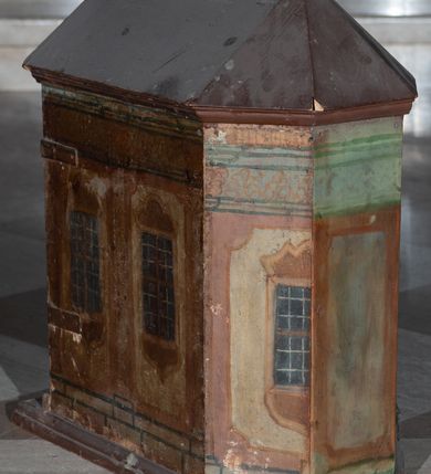 Zdjęcie nr 5: Tabernakulum w formie drewnianej skrzynki, zamkniętej trójbocznie, imitującej budynek, przykrytej wielospadowym dachem, na profilowanym cokole. Drzwiczki dwuskrzydłowe, w kształcie stojącego prostokąta, zwieńczone trójkątnym szczytem, zamocowane na zawiasach z okuciami. Dekorowane malowanymi płycinami o fantazyjnym kształcie i ornamentem.  Ścianki boczne z malowaną architekturą w kolorze jasnobrązowym, dwuosiowe, wydzielone pilastrami dźwigającymi belkowanie. Na osiach malowane okienka w dekoracyjnych opaskach. Na tylnej ściance namalowane belkowanie oraz dwie połówki okien w dekoracyjnych opaskach. Wnętrze tabernakulum polichromowane w kolorze jasnoniebieskim ze złoconymi gwiazdami. Drzwiczki po wewnętrznej stronie z malowaną dekoracją w postaci cokołów z wazonami i kwiatami, na jasnoniebieskim tle, w górnej partii ornament. 
