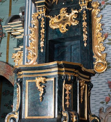 Zdjęcie nr 5: Ambona przyścienna, wisząca, z dostępem przez drzwi w zaplecku, od strony zakrystii. Kosz na planie kwadratu o wklęsłych ściankach i ściętych narożach, na podstawie w postaci wałka dekorowanego rocaille’m, z podwieszeniem w formie zbliżonej do stożka zakończonego szyszką, w którego ściętych narożach znajdują się spływy wolutowe dekorowane kampanulami i pola ozdobione rocaille’m. Kosz w narożach ujęty pilastrami o trzonach przechodzących w spływy wolutowe, dekorowanych kampanulami i rocaille’m, dźwigających niepełne belkowanie stanowiące parapet. Ścianki kosza zdobione płycinami z rocaille’m. Zaplecek ujęty pilastrami o kanelowanych trzonach, zakończonymi konsolami z podwieszonymi kampanulami, dźwigającymi imposty oraz fragmenty, przerwanego, wolutowego przyczółka. Pomiędzy nimi, w polu zwieńczenia gołębica Ducha Świętego w otoku z obłoków, w glorii promienistej. W polu zaplecka drzwi, ponad nimi rocaille. Zaplecek ujęty uszami utworzonymi ze spływów wolutowych, liści palmowych i kwiatów. Struktura polichromowana na sposób imitujący marmoryzację w kolorze ciemnej zieleni ze złoconymi profilami i ornamentyką, gołębica srebrzona. 