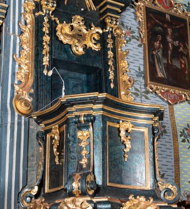 Zdjęcie nr 4: Ambona przyścienna, wisząca, z dostępem przez drzwi w zaplecku, od strony zakrystii. Kosz na planie kwadratu o wklęsłych ściankach i ściętych narożach, na podstawie w postaci wałka dekorowanego rocaille’m, z podwieszeniem w formie zbliżonej do stożka zakończonego szyszką, w którego ściętych narożach znajdują się spływy wolutowe dekorowane kampanulami i pola ozdobione rocaille’m. Kosz w narożach ujęty pilastrami o trzonach przechodzących w spływy wolutowe, dekorowanych kampanulami i rocaille’m, dźwigających niepełne belkowanie stanowiące parapet. Ścianki kosza zdobione płycinami z rocaille’m. Zaplecek ujęty pilastrami o kanelowanych trzonach, zakończonymi konsolami z podwieszonymi kampanulami, dźwigającymi imposty oraz fragmenty, przerwanego, wolutowego przyczółka. Pomiędzy nimi, w polu zwieńczenia gołębica Ducha Świętego w otoku z obłoków, w glorii promienistej. W polu zaplecka drzwi, ponad nimi rocaille. Zaplecek ujęty uszami utworzonymi ze spływów wolutowych, liści palmowych i kwiatów. Struktura polichromowana na sposób imitujący marmoryzację w kolorze ciemnej zieleni ze złoconymi profilami i ornamentyką, gołębica srebrzona. 