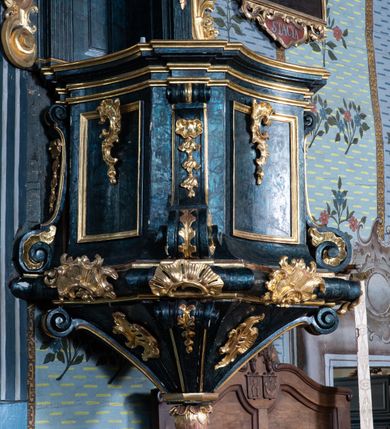 Zdjęcie nr 7: Ambona przyścienna, wisząca, z dostępem przez drzwi w zaplecku, od strony zakrystii. Kosz na planie kwadratu o wklęsłych ściankach i ściętych narożach, na podstawie w postaci wałka dekorowanego rocaille’m, z podwieszeniem w formie zbliżonej do stożka zakończonego szyszką, w którego ściętych narożach znajdują się spływy wolutowe dekorowane kampanulami i pola ozdobione rocaille’m. Kosz w narożach ujęty pilastrami o trzonach przechodzących w spływy wolutowe, dekorowanych kampanulami i rocaille’m, dźwigających niepełne belkowanie stanowiące parapet. Ścianki kosza zdobione płycinami z rocaille’m. Zaplecek ujęty pilastrami o kanelowanych trzonach, zakończonymi konsolami z podwieszonymi kampanulami, dźwigającymi imposty oraz fragmenty, przerwanego, wolutowego przyczółka. Pomiędzy nimi, w polu zwieńczenia gołębica Ducha Świętego w otoku z obłoków, w glorii promienistej. W polu zaplecka drzwi, ponad nimi rocaille. Zaplecek ujęty uszami utworzonymi ze spływów wolutowych, liści palmowych i kwiatów. Struktura polichromowana na sposób imitujący marmoryzację w kolorze ciemnej zieleni ze złoconymi profilami i ornamentyką, gołębica srebrzona. 