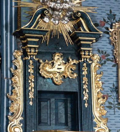 Zdjęcie nr 9: Ambona przyścienna, wisząca, z dostępem przez drzwi w zaplecku, od strony zakrystii. Kosz na planie kwadratu o wklęsłych ściankach i ściętych narożach, na podstawie w postaci wałka dekorowanego rocaille’m, z podwieszeniem w formie zbliżonej do stożka zakończonego szyszką, w którego ściętych narożach znajdują się spływy wolutowe dekorowane kampanulami i pola ozdobione rocaille’m. Kosz w narożach ujęty pilastrami o trzonach przechodzących w spływy wolutowe, dekorowanych kampanulami i rocaille’m, dźwigających niepełne belkowanie stanowiące parapet. Ścianki kosza zdobione płycinami z rocaille’m. Zaplecek ujęty pilastrami o kanelowanych trzonach, zakończonymi konsolami z podwieszonymi kampanulami, dźwigającymi imposty oraz fragmenty, przerwanego, wolutowego przyczółka. Pomiędzy nimi, w polu zwieńczenia gołębica Ducha Świętego w otoku z obłoków, w glorii promienistej. W polu zaplecka drzwi, ponad nimi rocaille. Zaplecek ujęty uszami utworzonymi ze spływów wolutowych, liści palmowych i kwiatów. Struktura polichromowana na sposób imitujący marmoryzację w kolorze ciemnej zieleni ze złoconymi profilami i ornamentyką, gołębica srebrzona. 