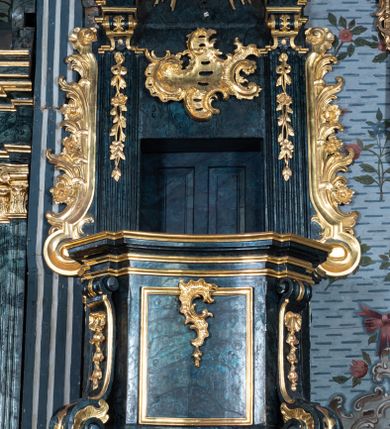 Zdjęcie nr 2: Ambona przyścienna, wisząca, z dostępem przez drzwi w zaplecku, od strony zakrystii. Kosz na planie kwadratu o wklęsłych ściankach i ściętych narożach, na podstawie w postaci wałka dekorowanego rocaille’m, z podwieszeniem w formie zbliżonej do stożka zakończonego szyszką, w którego ściętych narożach znajdują się spływy wolutowe dekorowane kampanulami i pola ozdobione rocaille’m. Kosz w narożach ujęty pilastrami o trzonach przechodzących w spływy wolutowe, dekorowanych kampanulami i rocaille’m, dźwigających niepełne belkowanie stanowiące parapet. Ścianki kosza zdobione płycinami z rocaille’m. Zaplecek ujęty pilastrami o kanelowanych trzonach, zakończonymi konsolami z podwieszonymi kampanulami, dźwigającymi imposty oraz fragmenty, przerwanego, wolutowego przyczółka. Pomiędzy nimi, w polu zwieńczenia gołębica Ducha Świętego w otoku z obłoków, w glorii promienistej. W polu zaplecka drzwi, ponad nimi rocaille. Zaplecek ujęty uszami utworzonymi ze spływów wolutowych, liści palmowych i kwiatów. Struktura polichromowana na sposób imitujący marmoryzację w kolorze ciemnej zieleni ze złoconymi profilami i ornamentyką, gołębica srebrzona. 