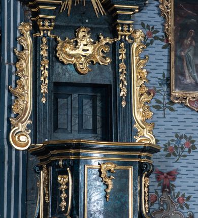 Zdjęcie nr 8: Ambona przyścienna, wisząca, z dostępem przez drzwi w zaplecku, od strony zakrystii. Kosz na planie kwadratu o wklęsłych ściankach i ściętych narożach, na podstawie w postaci wałka dekorowanego rocaille’m, z podwieszeniem w formie zbliżonej do stożka zakończonego szyszką, w którego ściętych narożach znajdują się spływy wolutowe dekorowane kampanulami i pola ozdobione rocaille’m. Kosz w narożach ujęty pilastrami o trzonach przechodzących w spływy wolutowe, dekorowanych kampanulami i rocaille’m, dźwigających niepełne belkowanie stanowiące parapet. Ścianki kosza zdobione płycinami z rocaille’m. Zaplecek ujęty pilastrami o kanelowanych trzonach, zakończonymi konsolami z podwieszonymi kampanulami, dźwigającymi imposty oraz fragmenty, przerwanego, wolutowego przyczółka. Pomiędzy nimi, w polu zwieńczenia gołębica Ducha Świętego w otoku z obłoków, w glorii promienistej. W polu zaplecka drzwi, ponad nimi rocaille. Zaplecek ujęty uszami utworzonymi ze spływów wolutowych, liści palmowych i kwiatów. Struktura polichromowana na sposób imitujący marmoryzację w kolorze ciemnej zieleni ze złoconymi profilami i ornamentyką, gołębica srebrzona. 