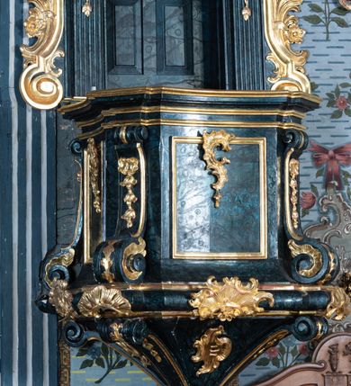 Zdjęcie nr 6: Ambona przyścienna, wisząca, z dostępem przez drzwi w zaplecku, od strony zakrystii. Kosz na planie kwadratu o wklęsłych ściankach i ściętych narożach, na podstawie w postaci wałka dekorowanego rocaille’m, z podwieszeniem w formie zbliżonej do stożka zakończonego szyszką, w którego ściętych narożach znajdują się spływy wolutowe dekorowane kampanulami i pola ozdobione rocaille’m. Kosz w narożach ujęty pilastrami o trzonach przechodzących w spływy wolutowe, dekorowanych kampanulami i rocaille’m, dźwigających niepełne belkowanie stanowiące parapet. Ścianki kosza zdobione płycinami z rocaille’m. Zaplecek ujęty pilastrami o kanelowanych trzonach, zakończonymi konsolami z podwieszonymi kampanulami, dźwigającymi imposty oraz fragmenty, przerwanego, wolutowego przyczółka. Pomiędzy nimi, w polu zwieńczenia gołębica Ducha Świętego w otoku z obłoków, w glorii promienistej. W polu zaplecka drzwi, ponad nimi rocaille. Zaplecek ujęty uszami utworzonymi ze spływów wolutowych, liści palmowych i kwiatów. Struktura polichromowana na sposób imitujący marmoryzację w kolorze ciemnej zieleni ze złoconymi profilami i ornamentyką, gołębica srebrzona. 