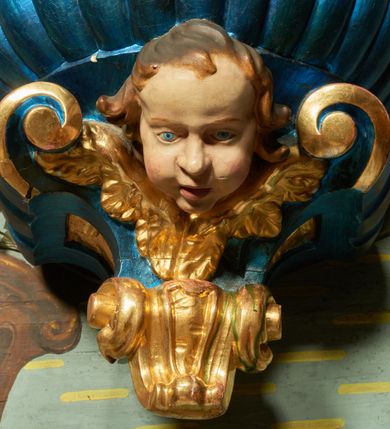 Zdjęcie nr 2: Chrzcielnica przyścienna. Czara półkolista z imitacją puklowania, z wywiniętym brzegiem, podtrzymywana u dołu przez dwie konsole, zakończone wolutą z uskrzydloną główką anielską. Nakryta płaską pokrywą na planie ściętego koła, na której znajduje się niewielka grupa rzeźbiarska Chrztu Chrystusa. Na pokrywie dwa rzędy ornamentalnych cęg, dolny zwrócony do wewnątrz, górny na zewnątrz, tworzące kabłąki podtrzymujące gołębicę Ducha Świętego w otoku z obłoków i glorii promienistej. Chrzcielnica i pokrywa laserowane w kolorze niebieskim, detale złocone i srebrzone; główka anielska i grupa rzeźbiarska polichromowane naturalistycznie. 