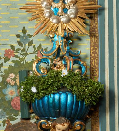 Zdjęcie nr 1: Chrzcielnica przyścienna. Czara półkolista z imitacją puklowania, z wywiniętym brzegiem, podtrzymywana u dołu przez dwie konsole, zakończone wolutą z uskrzydloną główką anielską. Nakryta płaską pokrywą na planie ściętego koła, na której znajduje się niewielka grupa rzeźbiarska Chrztu Chrystusa. Na pokrywie dwa rzędy ornamentalnych cęg, dolny zwrócony do wewnątrz, górny na zewnątrz, tworzące kabłąki podtrzymujące gołębicę Ducha Świętego w otoku z obłoków i glorii promienistej. Chrzcielnica i pokrywa laserowane w kolorze niebieskim, detale złocone i srebrzone; główka anielska i grupa rzeźbiarska polichromowane naturalistycznie. 