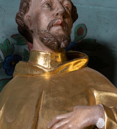 Zdjęcie nr 6: Rzeźba ścięta z tyłu, drążona, ustawiona na niskiej podstawie o ściętych narożach. Święty ukazany frontalnie, z głową skierowaną w lewo, w kontrapoście. W prawej, ugiętej i lekko uniesionej ręce trzyma krucyfiks, lewą dłoń kładzie na piersi. Twarz ma szczupłą, ascetyczną, o podkreślonej linii ściągniętych łuków brwiowych, głęboko osadzonych oczach z wyraźnie rytą górną powieką, skierowanych ku górze, dużym nosie i pełnych ustach z szeroką górną wargą. Twarz okolona krótką kręconą brodą oraz krótkimi falistymi puklami, na głowie tonsura. Ubrany jest w ściśle przylegający do ciała habit, przewiązany w talii, z lekko marszczonym szkaplerzem, na ramiona i plecy ma narzuconą pelerynkę z kapturem, z przodu krótką, a z tyłu sięgającą ziemi. Polichromia ciała naturalistyczna, szaty i atrybuty złocone.