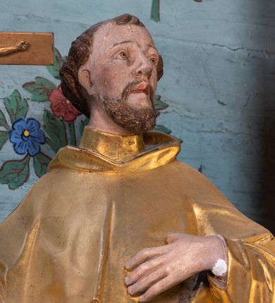 Zdjęcie nr 5: Rzeźba ścięta z tyłu, drążona, ustawiona na niskiej podstawie o ściętych narożach. Święty ukazany frontalnie, z głową skierowaną w lewo, w kontrapoście. W prawej, ugiętej i lekko uniesionej ręce trzyma krucyfiks, lewą dłoń kładzie na piersi. Twarz ma szczupłą, ascetyczną, o podkreślonej linii ściągniętych łuków brwiowych, głęboko osadzonych oczach z wyraźnie rytą górną powieką, skierowanych ku górze, dużym nosie i pełnych ustach z szeroką górną wargą. Twarz okolona krótką kręconą brodą oraz krótkimi falistymi puklami, na głowie tonsura. Ubrany jest w ściśle przylegający do ciała habit, przewiązany w talii, z lekko marszczonym szkaplerzem, na ramiona i plecy ma narzuconą pelerynkę z kapturem, z przodu krótką, a z tyłu sięgającą ziemi. Polichromia ciała naturalistyczna, szaty i atrybuty złocone.