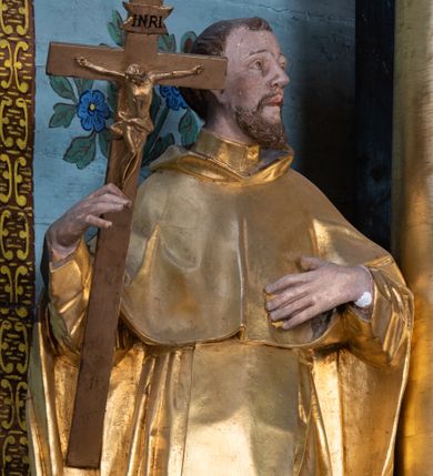 Zdjęcie nr 1: Rzeźba ścięta z tyłu, drążona, ustawiona na niskiej podstawie o ściętych narożach. Święty ukazany frontalnie, z głową skierowaną w lewo, w kontrapoście. W prawej, ugiętej i lekko uniesionej ręce trzyma krucyfiks, lewą dłoń kładzie na piersi. Twarz ma szczupłą, ascetyczną, o podkreślonej linii ściągniętych łuków brwiowych, głęboko osadzonych oczach z wyraźnie rytą górną powieką, skierowanych ku górze, dużym nosie i pełnych ustach z szeroką górną wargą. Twarz okolona krótką kręconą brodą oraz krótkimi falistymi puklami, na głowie tonsura. Ubrany jest w ściśle przylegający do ciała habit, przewiązany w talii, z lekko marszczonym szkaplerzem, na ramiona i plecy ma narzuconą pelerynkę z kapturem, z przodu krótką, a z tyłu sięgającą ziemi. Polichromia ciała naturalistyczna, szaty i atrybuty złocone.