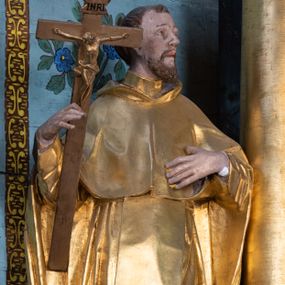 Zdjęcie nr 1: Rzeźba ścięta z tyłu, drążona, ustawiona na niskiej podstawie o ściętych narożach. Święty ukazany frontalnie, z głową skierowaną w lewo, w kontrapoście. W prawej, ugiętej i lekko uniesionej ręce trzyma krucyfiks, lewą dłoń kładzie na piersi. Twarz ma szczupłą, ascetyczną, o podkreślonej linii ściągniętych łuków brwiowych, głęboko osadzonych oczach z wyraźnie rytą górną powieką, skierowanych ku górze, dużym nosie i pełnych ustach z szeroką górną wargą. Twarz okolona krótką kręconą brodą oraz krótkimi falistymi puklami, na głowie tonsura. Ubrany jest w ściśle przylegający do ciała habit, przewiązany w talii, z lekko marszczonym szkaplerzem, na ramiona i plecy ma narzuconą pelerynkę z kapturem, z przodu krótką, a z tyłu sięgającą ziemi. Polichromia ciała naturalistyczna, szaty i atrybuty złocone.