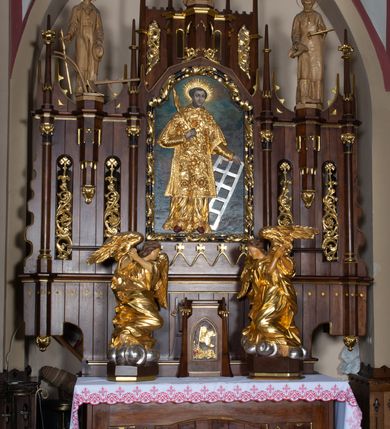 Zdjęcie nr 13: Nastawa architektoniczna, trójosiowa, jednokondygnacyjna ze zwieńczeniem, z predellą. Ołtarz prostopadłościenny, z drewnianą mensą i kamiennym portatylem. Antepedium ujęte grubą opaską, w polu płaskorzeźbiona otwarta księga z napisem „UBODZY / SKARBEM / KOŚCIOŁA” z liściem palmy i kratą. Na mensie tabernakulum w formie drewnianej skrzynki flankowanej pinaklami z kwiatonami. Drzwiczki w kształcie prostokąta zamkniętego łukiem ostrym, w ramie, z przedstawieniem baranka apokaliptycznego. Tabernakulum flankowane dwiema figurami klęczących na obłokach aniołów.  Osie nastawy wydzielone wąskimi kolumnami dźwigającymi stylizowane pinakle. W polu środkowym wnęka w formie stojącego prostokąta zamkniętego łukiem odcinkowym nadwieszonym, z obrazem św. Wawrzyńca, ujętym ramą zdobioną ornamentem roślinnym. W polach osi bocznych po dwie ażurowe stylizowane blendy wypełnione ornamentem roślinnym, pomiędzy nimi, w górnej partii, ośmioboczne wsporniki, na których stoją figury św. Izydora i św. Kryspina. Zwieńczenie w formie ośmiobocznego cokołu z figurą św. Józefa, flankowanego dwiema parami pinakli i blendami z dekoracja roślinną. Struktura pokryta politurą w kolorze ciemnobrązowym, ornamentyka i detale architektoniczne złocone. 
