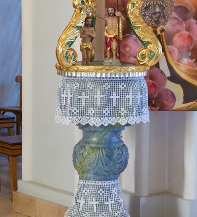 Zdjęcie nr 1: Chrzcielnica w typie kielichowym, stojąca. Stopa w kształcie ćwierćwałka przechodząca w zwężającą się tuleję. Trzon prosty, ujęty dwoma wklęsłymi pierścieniami. Nodus w formie spłaszczonej kuli, zdobionej motywem liści akantu. Czara półkolista, dekorowana wzorem imitującym puklowanie, z profilowanym brzegiem. 
Pokrywa na planie koła, w formie podstawy, której krawędź dekorowana jest motywem wieńca laurowego. Na niej znajdują się trzy kabłąki w formie spływów wolutowych zdobionych liśćmi akantu, zakończone trzema mniejszymi, stykającymi się wolutami zdobionymi liśćmi akantu. Podtrzymują one cokolik dekorowany lambrekinem z chwostami, na którym osadzona jest korona w typie zamkniętym, zwieńczona krzyżykiem. Na pokrywie, pośrodku ustawiona jest rzeźbiona grupa przedstawiająca Chrzest Chrystusa w Jordanie, z figurami zanurzonego w wodzie Chrystusa i stojącego na skale św. Jana Chrzciciela. Nad nimi, na złączeniu wolut, unosi się gołębica Ducha Świetego na tle glorii promienistej. 
Chrzcielnica polichromowana na sposób imitujący marmoryzację w odcieniach niebieskiego z żółtym żyłkowaniem; pokrywa złocona, srebrzona, zdobiona laserunkami, polichromowana. Polichromia ciał Chrystusa i św. Jana Chrzciciela naturalistyczna.