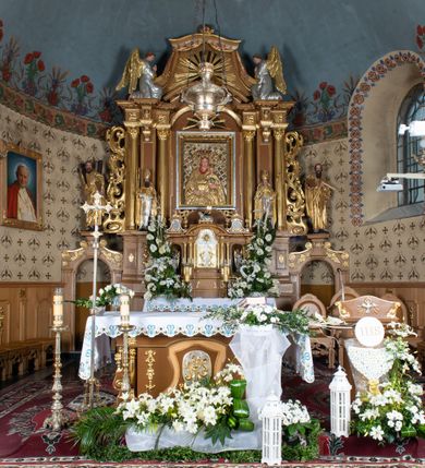 Zdjęcie nr 4: Nastawa ołtarzowa architektoniczna, jednoosiowa, jednokondygnacyjna z bramkami i zwieńczeniem, ustawiona na wysokim, dwustrefowym cokole. Ołtarz prostopadłościenny, murowany, umieszczony w drewnianej obudowie. Antependium ujęte ramą dekorowaną plecionką, podzielone na trzy pola: w środkowym płaskorzeźbione przedstawienie Ostatniej Wieczerzy według Leonarda da Vinci, a po bokach ornament roślinny o wertykalnym układzie.

Tabernakulum architektoniczne, jednokondygnacyjne, trójosiowe, ustawione na zdwojonym na cokole z dwoma zwieńczeniami na osiach skrajnych. Osie wydzielone są parami kolumn o kapitelach kompozytowych, oś środkowa wysunięta do przodu. Podpory dźwigają pełne belkowanie wyłamane na ich linii, przerwane nad polem środkowym, gdzie kolumny dźwigają przerwany półkolisty przyczółek połączony z łukiem gzymsu, nad nim ustawiona jest rzeźba Baranka leżącego na księdze z siedmioma pieczęciami. W polu środkowym dwuskrzydłowe drzwiczki do metalowego tabernakulum ujęte ramą w formie stojącego prostokąta, zamkniętego łukiem nadwieszonym z długim uskokiem. Na drzwiczkach znajdują się dwa aplikowane klęczące anioły, powyżej Chrismo w medalionie, otoczonym glorią. W osiach bocznych są nisze zamknięte konchami z figurkami aniołów, zamknięte zwieńczeniami w formie stojących prostokątów z falistym gzymsem i gałązkami lilii na szczycie; w polu srebrzone, owalne medaliony. Po bokach kondygnacji na wolutowych uszach oraz po bokach zwieńczeń znajdują się wazony ze słonecznikami. Cokół zdobiony jest ornamentem akantowym.

Kondygnacja ujęta jest dwiema parami kolumn o kapitelach kompozytowych oraz stojącą między nimi parą filarów. Podpory dźwigają pełne belkowanie o silnie wysuniętym gzymsie, wyłamane na osiach bocznych, z podwieszonymi szyszkami na tle filarów; przerwane nad polem środkowym. W centrum pole w formie stojącego prostokąta ujęte ramą dekorowaną ornamentem roślinnym z płaskorzeźbionym przedstawieniem Matki Boskiej z Dzieciątkiem. Na zasuwie obrazy: „Najświętsze Serce Pana Jezusa”, „Zwiastowanie” oraz „Św. Mikołaj”. Nad bramkami oraz pomiędzy kolumnami ustawione są rzeźby. Od lewej kolejno: św. Andrzej, św. Wojciech, św. Stanisław i na końcu po prawej św. Tomasz. Nad polem głównym znajduje się przeszklony prześwit w formie półkola z rzeźbioną gołębicą Ducha Świętego na tle glorii. Powyżej pole zamknięte półkoliście fragmentem gzymsu z rzeźbionym popiersiem błogosławiącego Boga Ojca pośród obłoków. Zwieńczenie w formie zbliżonej do stojącego prostokąta o wklęsłych bokach z gzymsem i z podwieszonym lambrekinem oraz kotarami spływającymi na boki. W polu przeszklony prześwit w formie kwadratu zamkniętego łukiem nadwieszonym z rzeźbionym monogramem Marii na tle glorii. Po bokach zwieńczenia ustawione są dwie duże figury klęczących aniołów.

Bramki po bokach ołtarza ustawione są na wysokości cokołu nastawy; przejścia ujęte są parą kanelowanych pilastrów, zamknięte łukiem utworzonym z dwóch przeciwstawnych wolut wspierających wieńczące cokoły. Przejścia w formie wydłużonego stojącego prostokąta zamkniętego łukiem półkolistym nadwieszonym.

Całość ołtarza oprócz tabernakulum dekorowana jest ornamentem małżowinowo-chrząstkowym. Uszy flankujące kondygnację utworzone są również z ornamentu małżowinowo-chrząstkowego. Na zamknięciu zwieńczenia znajdują się dwie uskrzydlone główki anielskie skierowane na wprost z podwieszonymi u szyi chustami. Struktura ołtarza polichromowana jest w kolorze brązowym ze złoconymi kolumnami, ornamentyką i profilami, detale srebrzone.