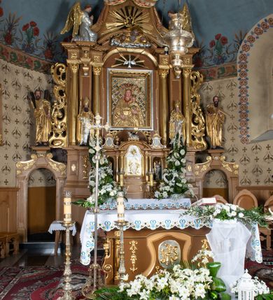 Zdjęcie nr 3: Nastawa ołtarzowa architektoniczna, jednoosiowa, jednokondygnacyjna z bramkami i zwieńczeniem, ustawiona na wysokim, dwustrefowym cokole. Ołtarz prostopadłościenny, murowany, umieszczony w drewnianej obudowie. Antependium ujęte ramą dekorowaną plecionką, podzielone na trzy pola: w środkowym płaskorzeźbione przedstawienie Ostatniej Wieczerzy według Leonarda da Vinci, a po bokach ornament roślinny o wertykalnym układzie.

Tabernakulum architektoniczne, jednokondygnacyjne, trójosiowe, ustawione na zdwojonym na cokole z dwoma zwieńczeniami na osiach skrajnych. Osie wydzielone są parami kolumn o kapitelach kompozytowych, oś środkowa wysunięta do przodu. Podpory dźwigają pełne belkowanie wyłamane na ich linii, przerwane nad polem środkowym, gdzie kolumny dźwigają przerwany półkolisty przyczółek połączony z łukiem gzymsu, nad nim ustawiona jest rzeźba Baranka leżącego na księdze z siedmioma pieczęciami. W polu środkowym dwuskrzydłowe drzwiczki do metalowego tabernakulum ujęte ramą w formie stojącego prostokąta, zamkniętego łukiem nadwieszonym z długim uskokiem. Na drzwiczkach znajdują się dwa aplikowane klęczące anioły, powyżej Chrismo w medalionie, otoczonym glorią. W osiach bocznych są nisze zamknięte konchami z figurkami aniołów, zamknięte zwieńczeniami w formie stojących prostokątów z falistym gzymsem i gałązkami lilii na szczycie; w polu srebrzone, owalne medaliony. Po bokach kondygnacji na wolutowych uszach oraz po bokach zwieńczeń znajdują się wazony ze słonecznikami. Cokół zdobiony jest ornamentem akantowym.

Kondygnacja ujęta jest dwiema parami kolumn o kapitelach kompozytowych oraz stojącą między nimi parą filarów. Podpory dźwigają pełne belkowanie o silnie wysuniętym gzymsie, wyłamane na osiach bocznych, z podwieszonymi szyszkami na tle filarów; przerwane nad polem środkowym. W centrum pole w formie stojącego prostokąta ujęte ramą dekorowaną ornamentem roślinnym z płaskorzeźbionym przedstawieniem Matki Boskiej z Dzieciątkiem. Na zasuwie obrazy: „Najświętsze Serce Pana Jezusa”, „Zwiastowanie” oraz „Św. Mikołaj”. Nad bramkami oraz pomiędzy kolumnami ustawione są rzeźby. Od lewej kolejno: św. Andrzej, św. Wojciech, św. Stanisław i na końcu po prawej św. Tomasz. Nad polem głównym znajduje się przeszklony prześwit w formie półkola z rzeźbioną gołębicą Ducha Świętego na tle glorii. Powyżej pole zamknięte półkoliście fragmentem gzymsu z rzeźbionym popiersiem błogosławiącego Boga Ojca pośród obłoków. Zwieńczenie w formie zbliżonej do stojącego prostokąta o wklęsłych bokach z gzymsem i z podwieszonym lambrekinem oraz kotarami spływającymi na boki. W polu przeszklony prześwit w formie kwadratu zamkniętego łukiem nadwieszonym z rzeźbionym monogramem Marii na tle glorii. Po bokach zwieńczenia ustawione są dwie duże figury klęczących aniołów.

Bramki po bokach ołtarza ustawione są na wysokości cokołu nastawy; przejścia ujęte są parą kanelowanych pilastrów, zamknięte łukiem utworzonym z dwóch przeciwstawnych wolut wspierających wieńczące cokoły. Przejścia w formie wydłużonego stojącego prostokąta zamkniętego łukiem półkolistym nadwieszonym.

Całość ołtarza oprócz tabernakulum dekorowana jest ornamentem małżowinowo-chrząstkowym. Uszy flankujące kondygnację utworzone są również z ornamentu małżowinowo-chrząstkowego. Na zamknięciu zwieńczenia znajdują się dwie uskrzydlone główki anielskie skierowane na wprost z podwieszonymi u szyi chustami. Struktura ołtarza polichromowana jest w kolorze brązowym ze złoconymi kolumnami, ornamentyką i profilami, detale srebrzone.