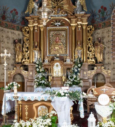 Zdjęcie nr 2: Nastawa ołtarzowa architektoniczna, jednoosiowa, jednokondygnacyjna z bramkami i zwieńczeniem, ustawiona na wysokim, dwustrefowym cokole. Ołtarz prostopadłościenny, murowany, umieszczony w drewnianej obudowie. Antependium ujęte ramą dekorowaną plecionką, podzielone na trzy pola: w środkowym płaskorzeźbione przedstawienie Ostatniej Wieczerzy według Leonarda da Vinci, a po bokach ornament roślinny o wertykalnym układzie.

Tabernakulum architektoniczne, jednokondygnacyjne, trójosiowe, ustawione na zdwojonym na cokole z dwoma zwieńczeniami na osiach skrajnych. Osie wydzielone są parami kolumn o kapitelach kompozytowych, oś środkowa wysunięta do przodu. Podpory dźwigają pełne belkowanie wyłamane na ich linii, przerwane nad polem środkowym, gdzie kolumny dźwigają przerwany półkolisty przyczółek połączony z łukiem gzymsu, nad nim ustawiona jest rzeźba Baranka leżącego na księdze z siedmioma pieczęciami. W polu środkowym dwuskrzydłowe drzwiczki do metalowego tabernakulum ujęte ramą w formie stojącego prostokąta, zamkniętego łukiem nadwieszonym z długim uskokiem. Na drzwiczkach znajdują się dwa aplikowane klęczące anioły, powyżej Chrismo w medalionie, otoczonym glorią. W osiach bocznych są nisze zamknięte konchami z figurkami aniołów, zamknięte zwieńczeniami w formie stojących prostokątów z falistym gzymsem i gałązkami lilii na szczycie; w polu srebrzone, owalne medaliony. Po bokach kondygnacji na wolutowych uszach oraz po bokach zwieńczeń znajdują się wazony ze słonecznikami. Cokół zdobiony jest ornamentem akantowym.

Kondygnacja ujęta jest dwiema parami kolumn o kapitelach kompozytowych oraz stojącą między nimi parą filarów. Podpory dźwigają pełne belkowanie o silnie wysuniętym gzymsie, wyłamane na osiach bocznych, z podwieszonymi szyszkami na tle filarów; przerwane nad polem środkowym. W centrum pole w formie stojącego prostokąta ujęte ramą dekorowaną ornamentem roślinnym z płaskorzeźbionym przedstawieniem Matki Boskiej z Dzieciątkiem. Na zasuwie obrazy: „Najświętsze Serce Pana Jezusa”, „Zwiastowanie” oraz „Św. Mikołaj”. Nad bramkami oraz pomiędzy kolumnami ustawione są rzeźby. Od lewej kolejno: św. Andrzej, św. Wojciech, św. Stanisław i na końcu po prawej św. Tomasz. Nad polem głównym znajduje się przeszklony prześwit w formie półkola z rzeźbioną gołębicą Ducha Świętego na tle glorii. Powyżej pole zamknięte półkoliście fragmentem gzymsu z rzeźbionym popiersiem błogosławiącego Boga Ojca pośród obłoków. Zwieńczenie w formie zbliżonej do stojącego prostokąta o wklęsłych bokach z gzymsem i z podwieszonym lambrekinem oraz kotarami spływającymi na boki. W polu przeszklony prześwit w formie kwadratu zamkniętego łukiem nadwieszonym z rzeźbionym monogramem Marii na tle glorii. Po bokach zwieńczenia ustawione są dwie duże figury klęczących aniołów.

Bramki po bokach ołtarza ustawione są na wysokości cokołu nastawy; przejścia ujęte są parą kanelowanych pilastrów, zamknięte łukiem utworzonym z dwóch przeciwstawnych wolut wspierających wieńczące cokoły. Przejścia w formie wydłużonego stojącego prostokąta zamkniętego łukiem półkolistym nadwieszonym.

Całość ołtarza oprócz tabernakulum dekorowana jest ornamentem małżowinowo-chrząstkowym. Uszy flankujące kondygnację utworzone są również z ornamentu małżowinowo-chrząstkowego. Na zamknięciu zwieńczenia znajdują się dwie uskrzydlone główki anielskie skierowane na wprost z podwieszonymi u szyi chustami. Struktura ołtarza polichromowana jest w kolorze brązowym ze złoconymi kolumnami, ornamentyką i profilami, detale srebrzone.