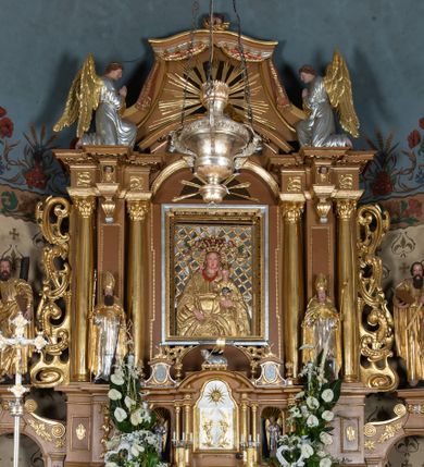 Zdjęcie nr 1: Nastawa ołtarzowa architektoniczna, jednoosiowa, jednokondygnacyjna z bramkami i zwieńczeniem, ustawiona na wysokim, dwustrefowym cokole. Ołtarz prostopadłościenny, murowany, umieszczony w drewnianej obudowie. Antependium ujęte ramą dekorowaną plecionką, podzielone na trzy pola: w środkowym płaskorzeźbione przedstawienie Ostatniej Wieczerzy według Leonarda da Vinci, a po bokach ornament roślinny o wertykalnym układzie.

Tabernakulum architektoniczne, jednokondygnacyjne, trójosiowe, ustawione na zdwojonym na cokole z dwoma zwieńczeniami na osiach skrajnych. Osie wydzielone są parami kolumn o kapitelach kompozytowych, oś środkowa wysunięta do przodu. Podpory dźwigają pełne belkowanie wyłamane na ich linii, przerwane nad polem środkowym, gdzie kolumny dźwigają przerwany półkolisty przyczółek połączony z łukiem gzymsu, nad nim ustawiona jest rzeźba Baranka leżącego na księdze z siedmioma pieczęciami. W polu środkowym dwuskrzydłowe drzwiczki do metalowego tabernakulum ujęte ramą w formie stojącego prostokąta, zamkniętego łukiem nadwieszonym z długim uskokiem. Na drzwiczkach znajdują się dwa aplikowane klęczące anioły, powyżej Chrismo w medalionie, otoczonym glorią. W osiach bocznych są nisze zamknięte konchami z figurkami aniołów, zamknięte zwieńczeniami w formie stojących prostokątów z falistym gzymsem i gałązkami lilii na szczycie; w polu srebrzone, owalne medaliony. Po bokach kondygnacji na wolutowych uszach oraz po bokach zwieńczeń znajdują się wazony ze słonecznikami. Cokół zdobiony jest ornamentem akantowym.

Kondygnacja ujęta jest dwiema parami kolumn o kapitelach kompozytowych oraz stojącą między nimi parą filarów. Podpory dźwigają pełne belkowanie o silnie wysuniętym gzymsie, wyłamane na osiach bocznych, z podwieszonymi szyszkami na tle filarów; przerwane nad polem środkowym. W centrum pole w formie stojącego prostokąta ujęte ramą dekorowaną ornamentem roślinnym z płaskorzeźbionym przedstawieniem Matki Boskiej z Dzieciątkiem. Na zasuwie obrazy: „Najświętsze Serce Pana Jezusa”, „Zwiastowanie” oraz „Św. Mikołaj”. Nad bramkami oraz pomiędzy kolumnami ustawione są rzeźby. Od lewej kolejno: św. Andrzej, św. Wojciech, św. Stanisław i na końcu po prawej św. Tomasz. Nad polem głównym znajduje się przeszklony prześwit w formie półkola z rzeźbioną gołębicą Ducha Świętego na tle glorii. Powyżej pole zamknięte półkoliście fragmentem gzymsu z rzeźbionym popiersiem błogosławiącego Boga Ojca pośród obłoków. Zwieńczenie w formie zbliżonej do stojącego prostokąta o wklęsłych bokach z gzymsem i z podwieszonym lambrekinem oraz kotarami spływającymi na boki. W polu przeszklony prześwit w formie kwadratu zamkniętego łukiem nadwieszonym z rzeźbionym monogramem Marii na tle glorii. Po bokach zwieńczenia ustawione są dwie duże figury klęczących aniołów.

Bramki po bokach ołtarza ustawione są na wysokości cokołu nastawy; przejścia ujęte są parą kanelowanych pilastrów, zamknięte łukiem utworzonym z dwóch przeciwstawnych wolut wspierających wieńczące cokoły. Przejścia w formie wydłużonego stojącego prostokąta zamkniętego łukiem półkolistym nadwieszonym.

Całość ołtarza oprócz tabernakulum dekorowana jest ornamentem małżowinowo-chrząstkowym. Uszy flankujące kondygnację utworzone są również z ornamentu małżowinowo-chrząstkowego. Na zamknięciu zwieńczenia znajdują się dwie uskrzydlone główki anielskie skierowane na wprost z podwieszonymi u szyi chustami. Struktura ołtarza polichromowana jest w kolorze brązowym ze złoconymi kolumnami, ornamentyką i profilami, detale srebrzone.