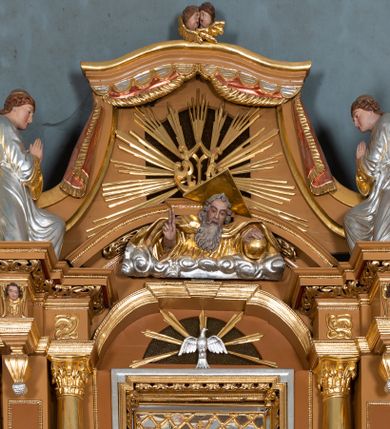 Zdjęcie nr 5: Nastawa ołtarzowa architektoniczna, jednoosiowa, jednokondygnacyjna z bramkami i zwieńczeniem, ustawiona na wysokim, dwustrefowym cokole. Ołtarz prostopadłościenny, murowany, umieszczony w drewnianej obudowie. Antependium ujęte ramą dekorowaną plecionką, podzielone na trzy pola: w środkowym płaskorzeźbione przedstawienie Ostatniej Wieczerzy według Leonarda da Vinci, a po bokach ornament roślinny o wertykalnym układzie.

Tabernakulum architektoniczne, jednokondygnacyjne, trójosiowe, ustawione na zdwojonym na cokole z dwoma zwieńczeniami na osiach skrajnych. Osie wydzielone są parami kolumn o kapitelach kompozytowych, oś środkowa wysunięta do przodu. Podpory dźwigają pełne belkowanie wyłamane na ich linii, przerwane nad polem środkowym, gdzie kolumny dźwigają przerwany półkolisty przyczółek połączony z łukiem gzymsu, nad nim ustawiona jest rzeźba Baranka leżącego na księdze z siedmioma pieczęciami. W polu środkowym dwuskrzydłowe drzwiczki do metalowego tabernakulum ujęte ramą w formie stojącego prostokąta, zamkniętego łukiem nadwieszonym z długim uskokiem. Na drzwiczkach znajdują się dwa aplikowane klęczące anioły, powyżej Chrismo w medalionie, otoczonym glorią. W osiach bocznych są nisze zamknięte konchami z figurkami aniołów, zamknięte zwieńczeniami w formie stojących prostokątów z falistym gzymsem i gałązkami lilii na szczycie; w polu srebrzone, owalne medaliony. Po bokach kondygnacji na wolutowych uszach oraz po bokach zwieńczeń znajdują się wazony ze słonecznikami. Cokół zdobiony jest ornamentem akantowym.

Kondygnacja ujęta jest dwiema parami kolumn o kapitelach kompozytowych oraz stojącą między nimi parą filarów. Podpory dźwigają pełne belkowanie o silnie wysuniętym gzymsie, wyłamane na osiach bocznych, z podwieszonymi szyszkami na tle filarów; przerwane nad polem środkowym. W centrum pole w formie stojącego prostokąta ujęte ramą dekorowaną ornamentem roślinnym z płaskorzeźbionym przedstawieniem Matki Boskiej z Dzieciątkiem. Na zasuwie obrazy: „Najświętsze Serce Pana Jezusa”, „Zwiastowanie” oraz „Św. Mikołaj”. Nad bramkami oraz pomiędzy kolumnami ustawione są rzeźby. Od lewej kolejno: św. Andrzej, św. Wojciech, św. Stanisław i na końcu po prawej św. Tomasz. Nad polem głównym znajduje się przeszklony prześwit w formie półkola z rzeźbioną gołębicą Ducha Świętego na tle glorii. Powyżej pole zamknięte półkoliście fragmentem gzymsu z rzeźbionym popiersiem błogosławiącego Boga Ojca pośród obłoków. Zwieńczenie w formie zbliżonej do stojącego prostokąta o wklęsłych bokach z gzymsem i z podwieszonym lambrekinem oraz kotarami spływającymi na boki. W polu przeszklony prześwit w formie kwadratu zamkniętego łukiem nadwieszonym z rzeźbionym monogramem Marii na tle glorii. Po bokach zwieńczenia ustawione są dwie duże figury klęczących aniołów.

Bramki po bokach ołtarza ustawione są na wysokości cokołu nastawy; przejścia ujęte są parą kanelowanych pilastrów, zamknięte łukiem utworzonym z dwóch przeciwstawnych wolut wspierających wieńczące cokoły. Przejścia w formie wydłużonego stojącego prostokąta zamkniętego łukiem półkolistym nadwieszonym.

Całość ołtarza oprócz tabernakulum dekorowana jest ornamentem małżowinowo-chrząstkowym. Uszy flankujące kondygnację utworzone są również z ornamentu małżowinowo-chrząstkowego. Na zamknięciu zwieńczenia znajdują się dwie uskrzydlone główki anielskie skierowane na wprost z podwieszonymi u szyi chustami. Struktura ołtarza polichromowana jest w kolorze brązowym ze złoconymi kolumnami, ornamentyką i profilami, detale srebrzone.