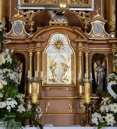 Zdjęcie nr 8: Nastawa ołtarzowa architektoniczna, jednoosiowa, jednokondygnacyjna z bramkami i zwieńczeniem, ustawiona na wysokim, dwustrefowym cokole. Ołtarz prostopadłościenny, murowany, umieszczony w drewnianej obudowie. Antependium ujęte ramą dekorowaną plecionką, podzielone na trzy pola: w środkowym płaskorzeźbione przedstawienie Ostatniej Wieczerzy według Leonarda da Vinci, a po bokach ornament roślinny o wertykalnym układzie.

Tabernakulum architektoniczne, jednokondygnacyjne, trójosiowe, ustawione na zdwojonym na cokole z dwoma zwieńczeniami na osiach skrajnych. Osie wydzielone są parami kolumn o kapitelach kompozytowych, oś środkowa wysunięta do przodu. Podpory dźwigają pełne belkowanie wyłamane na ich linii, przerwane nad polem środkowym, gdzie kolumny dźwigają przerwany półkolisty przyczółek połączony z łukiem gzymsu, nad nim ustawiona jest rzeźba Baranka leżącego na księdze z siedmioma pieczęciami. W polu środkowym dwuskrzydłowe drzwiczki do metalowego tabernakulum ujęte ramą w formie stojącego prostokąta, zamkniętego łukiem nadwieszonym z długim uskokiem. Na drzwiczkach znajdują się dwa aplikowane klęczące anioły, powyżej Chrismo w medalionie, otoczonym glorią. W osiach bocznych są nisze zamknięte konchami z figurkami aniołów, zamknięte zwieńczeniami w formie stojących prostokątów z falistym gzymsem i gałązkami lilii na szczycie; w polu srebrzone, owalne medaliony. Po bokach kondygnacji na wolutowych uszach oraz po bokach zwieńczeń znajdują się wazony ze słonecznikami. Cokół zdobiony jest ornamentem akantowym.

Kondygnacja ujęta jest dwiema parami kolumn o kapitelach kompozytowych oraz stojącą między nimi parą filarów. Podpory dźwigają pełne belkowanie o silnie wysuniętym gzymsie, wyłamane na osiach bocznych, z podwieszonymi szyszkami na tle filarów; przerwane nad polem środkowym. W centrum pole w formie stojącego prostokąta ujęte ramą dekorowaną ornamentem roślinnym z płaskorzeźbionym przedstawieniem Matki Boskiej z Dzieciątkiem. Na zasuwie obrazy: „Najświętsze Serce Pana Jezusa”, „Zwiastowanie” oraz „Św. Mikołaj”. Nad bramkami oraz pomiędzy kolumnami ustawione są rzeźby. Od lewej kolejno: św. Andrzej, św. Wojciech, św. Stanisław i na końcu po prawej św. Tomasz. Nad polem głównym znajduje się przeszklony prześwit w formie półkola z rzeźbioną gołębicą Ducha Świętego na tle glorii. Powyżej pole zamknięte półkoliście fragmentem gzymsu z rzeźbionym popiersiem błogosławiącego Boga Ojca pośród obłoków. Zwieńczenie w formie zbliżonej do stojącego prostokąta o wklęsłych bokach z gzymsem i z podwieszonym lambrekinem oraz kotarami spływającymi na boki. W polu przeszklony prześwit w formie kwadratu zamkniętego łukiem nadwieszonym z rzeźbionym monogramem Marii na tle glorii. Po bokach zwieńczenia ustawione są dwie duże figury klęczących aniołów.

Bramki po bokach ołtarza ustawione są na wysokości cokołu nastawy; przejścia ujęte są parą kanelowanych pilastrów, zamknięte łukiem utworzonym z dwóch przeciwstawnych wolut wspierających wieńczące cokoły. Przejścia w formie wydłużonego stojącego prostokąta zamkniętego łukiem półkolistym nadwieszonym.

Całość ołtarza oprócz tabernakulum dekorowana jest ornamentem małżowinowo-chrząstkowym. Uszy flankujące kondygnację utworzone są również z ornamentu małżowinowo-chrząstkowego. Na zamknięciu zwieńczenia znajdują się dwie uskrzydlone główki anielskie skierowane na wprost z podwieszonymi u szyi chustami. Struktura ołtarza polichromowana jest w kolorze brązowym ze złoconymi kolumnami, ornamentyką i profilami, detale srebrzone.