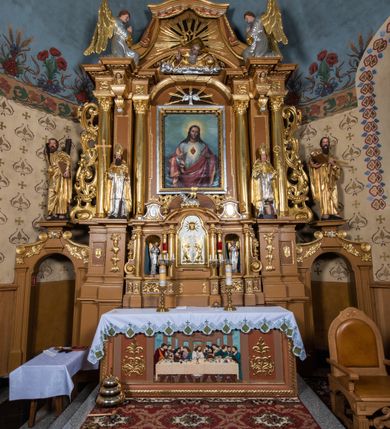 Zdjęcie nr 29: Nastawa ołtarzowa architektoniczna, jednoosiowa, jednokondygnacyjna z bramkami i zwieńczeniem, ustawiona na wysokim, dwustrefowym cokole. Ołtarz prostopadłościenny, murowany, umieszczony w drewnianej obudowie. Antependium ujęte ramą dekorowaną plecionką, podzielone na trzy pola: w środkowym płaskorzeźbione przedstawienie Ostatniej Wieczerzy według Leonarda da Vinci, a po bokach ornament roślinny o wertykalnym układzie.

Tabernakulum architektoniczne, jednokondygnacyjne, trójosiowe, ustawione na zdwojonym na cokole z dwoma zwieńczeniami na osiach skrajnych. Osie wydzielone są parami kolumn o kapitelach kompozytowych, oś środkowa wysunięta do przodu. Podpory dźwigają pełne belkowanie wyłamane na ich linii, przerwane nad polem środkowym, gdzie kolumny dźwigają przerwany półkolisty przyczółek połączony z łukiem gzymsu, nad nim ustawiona jest rzeźba Baranka leżącego na księdze z siedmioma pieczęciami. W polu środkowym dwuskrzydłowe drzwiczki do metalowego tabernakulum ujęte ramą w formie stojącego prostokąta, zamkniętego łukiem nadwieszonym z długim uskokiem. Na drzwiczkach znajdują się dwa aplikowane klęczące anioły, powyżej Chrismo w medalionie, otoczonym glorią. W osiach bocznych są nisze zamknięte konchami z figurkami aniołów, zamknięte zwieńczeniami w formie stojących prostokątów z falistym gzymsem i gałązkami lilii na szczycie; w polu srebrzone, owalne medaliony. Po bokach kondygnacji na wolutowych uszach oraz po bokach zwieńczeń znajdują się wazony ze słonecznikami. Cokół zdobiony jest ornamentem akantowym.

Kondygnacja ujęta jest dwiema parami kolumn o kapitelach kompozytowych oraz stojącą między nimi parą filarów. Podpory dźwigają pełne belkowanie o silnie wysuniętym gzymsie, wyłamane na osiach bocznych, z podwieszonymi szyszkami na tle filarów; przerwane nad polem środkowym. W centrum pole w formie stojącego prostokąta ujęte ramą dekorowaną ornamentem roślinnym z płaskorzeźbionym przedstawieniem Matki Boskiej z Dzieciątkiem. Na zasuwie obrazy: „Najświętsze Serce Pana Jezusa”, „Zwiastowanie” oraz „Św. Mikołaj”. Nad bramkami oraz pomiędzy kolumnami ustawione są rzeźby. Od lewej kolejno: św. Andrzej, św. Wojciech, św. Stanisław i na końcu po prawej św. Tomasz. Nad polem głównym znajduje się przeszklony prześwit w formie półkola z rzeźbioną gołębicą Ducha Świętego na tle glorii. Powyżej pole zamknięte półkoliście fragmentem gzymsu z rzeźbionym popiersiem błogosławiącego Boga Ojca pośród obłoków. Zwieńczenie w formie zbliżonej do stojącego prostokąta o wklęsłych bokach z gzymsem i z podwieszonym lambrekinem oraz kotarami spływającymi na boki. W polu przeszklony prześwit w formie kwadratu zamkniętego łukiem nadwieszonym z rzeźbionym monogramem Marii na tle glorii. Po bokach zwieńczenia ustawione są dwie duże figury klęczących aniołów.

Bramki po bokach ołtarza ustawione są na wysokości cokołu nastawy; przejścia ujęte są parą kanelowanych pilastrów, zamknięte łukiem utworzonym z dwóch przeciwstawnych wolut wspierających wieńczące cokoły. Przejścia w formie wydłużonego stojącego prostokąta zamkniętego łukiem półkolistym nadwieszonym.

Całość ołtarza oprócz tabernakulum dekorowana jest ornamentem małżowinowo-chrząstkowym. Uszy flankujące kondygnację utworzone są również z ornamentu małżowinowo-chrząstkowego. Na zamknięciu zwieńczenia znajdują się dwie uskrzydlone główki anielskie skierowane na wprost z podwieszonymi u szyi chustami. Struktura ołtarza polichromowana jest w kolorze brązowym ze złoconymi kolumnami, ornamentyką i profilami, detale srebrzone.