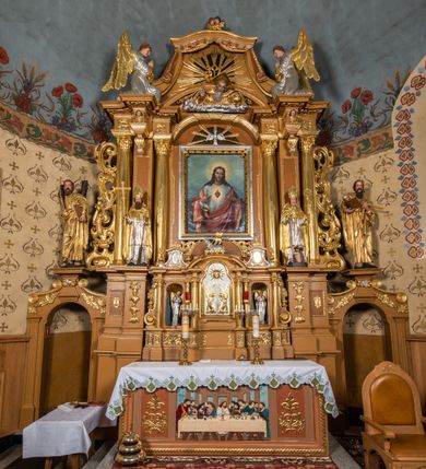 Zdjęcie nr 36: Nastawa ołtarzowa architektoniczna, jednoosiowa, jednokondygnacyjna z bramkami i zwieńczeniem, ustawiona na wysokim, dwustrefowym cokole. Ołtarz prostopadłościenny, murowany, umieszczony w drewnianej obudowie. Antependium ujęte ramą dekorowaną plecionką, podzielone na trzy pola: w środkowym płaskorzeźbione przedstawienie Ostatniej Wieczerzy według Leonarda da Vinci, a po bokach ornament roślinny o wertykalnym układzie.

Tabernakulum architektoniczne, jednokondygnacyjne, trójosiowe, ustawione na zdwojonym na cokole z dwoma zwieńczeniami na osiach skrajnych. Osie wydzielone są parami kolumn o kapitelach kompozytowych, oś środkowa wysunięta do przodu. Podpory dźwigają pełne belkowanie wyłamane na ich linii, przerwane nad polem środkowym, gdzie kolumny dźwigają przerwany półkolisty przyczółek połączony z łukiem gzymsu, nad nim ustawiona jest rzeźba Baranka leżącego na księdze z siedmioma pieczęciami. W polu środkowym dwuskrzydłowe drzwiczki do metalowego tabernakulum ujęte ramą w formie stojącego prostokąta, zamkniętego łukiem nadwieszonym z długim uskokiem. Na drzwiczkach znajdują się dwa aplikowane klęczące anioły, powyżej Chrismo w medalionie, otoczonym glorią. W osiach bocznych są nisze zamknięte konchami z figurkami aniołów, zamknięte zwieńczeniami w formie stojących prostokątów z falistym gzymsem i gałązkami lilii na szczycie; w polu srebrzone, owalne medaliony. Po bokach kondygnacji na wolutowych uszach oraz po bokach zwieńczeń znajdują się wazony ze słonecznikami. Cokół zdobiony jest ornamentem akantowym.

Kondygnacja ujęta jest dwiema parami kolumn o kapitelach kompozytowych oraz stojącą między nimi parą filarów. Podpory dźwigają pełne belkowanie o silnie wysuniętym gzymsie, wyłamane na osiach bocznych, z podwieszonymi szyszkami na tle filarów; przerwane nad polem środkowym. W centrum pole w formie stojącego prostokąta ujęte ramą dekorowaną ornamentem roślinnym z płaskorzeźbionym przedstawieniem Matki Boskiej z Dzieciątkiem. Na zasuwie obrazy: „Najświętsze Serce Pana Jezusa”, „Zwiastowanie” oraz „Św. Mikołaj”. Nad bramkami oraz pomiędzy kolumnami ustawione są rzeźby. Od lewej kolejno: św. Andrzej, św. Wojciech, św. Stanisław i na końcu po prawej św. Tomasz. Nad polem głównym znajduje się przeszklony prześwit w formie półkola z rzeźbioną gołębicą Ducha Świętego na tle glorii. Powyżej pole zamknięte półkoliście fragmentem gzymsu z rzeźbionym popiersiem błogosławiącego Boga Ojca pośród obłoków. Zwieńczenie w formie zbliżonej do stojącego prostokąta o wklęsłych bokach z gzymsem i z podwieszonym lambrekinem oraz kotarami spływającymi na boki. W polu przeszklony prześwit w formie kwadratu zamkniętego łukiem nadwieszonym z rzeźbionym monogramem Marii na tle glorii. Po bokach zwieńczenia ustawione są dwie duże figury klęczących aniołów.

Bramki po bokach ołtarza ustawione są na wysokości cokołu nastawy; przejścia ujęte są parą kanelowanych pilastrów, zamknięte łukiem utworzonym z dwóch przeciwstawnych wolut wspierających wieńczące cokoły. Przejścia w formie wydłużonego stojącego prostokąta zamkniętego łukiem półkolistym nadwieszonym.

Całość ołtarza oprócz tabernakulum dekorowana jest ornamentem małżowinowo-chrząstkowym. Uszy flankujące kondygnację utworzone są również z ornamentu małżowinowo-chrząstkowego. Na zamknięciu zwieńczenia znajdują się dwie uskrzydlone główki anielskie skierowane na wprost z podwieszonymi u szyi chustami. Struktura ołtarza polichromowana jest w kolorze brązowym ze złoconymi kolumnami, ornamentyką i profilami, detale srebrzone.