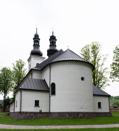 Zdjęcie nr 7: Kościół usytuowany jest w południowo-wschodniej części wsi, otoczony kamiennym murem, otynkowanym, krytym betonową płytą, w który wbudowano współczesne stacje drogi krzyżowej. Na plac kościelny prowadzą trzy bramy. Główna brama znajduje się w murze od strony zachodniej, nakryta jest ostrołukową arkadą. Dwie mniejsze bramy zlokalizowane są od strony południowej oraz od strony wschodniej. Na placu przed kościołem stoi pomnik z 1911 roku wystawiony na pamiątkę księdza Wojciecha Blaszyńskiego i działacza ludowego Stanisława Stojałowskiego. 
Świątynia jest orientowana, murowana, jednonawowa z węższym i krótkim prezbiterium, zamkniętym półkoliście oraz z wieżą na zakończeniu nawy. Do prezbiterium przylega zakrystia od północy i skarbczyk od południa. Od strony południowej dobudowana jest również niska kruchta. Wnętrze trójprzęsłowej nawy rozczłonkowane jest filarami przyściennymi wspierającymi sklepienie żagielkowe. Chór muzyczny trójarkadowy, wsparty na dwóch filarach. W ścianach korpusu nawowego znajdują się głębokie wnęki, w których umieszczone są prostokątne otwory okienne zamknięte półkolistym łukiem, po trzy od strony północnej i południowej; takie same okno znajduje się również w apsydzie od strony południowej oraz na chórze od strony zachodniej. Ponadto w apsydzie na wysokości zwieńczenia ołtarza głównego umieszczone jest małe, okrągłe okno. W kruchcie, skarbczyku i zakrystii okna są prostokątne. Drzwi główne dwuskrzydłowe, drewniane z metalowymi okuciami, zabezpieczone od wewnątrz dwiema żelaznymi zaporami umocowanymi w murze. Podobne drzwi znajdują się od strony południowej z kruchty. 
Fasada kościoła jest płaska, zakończona gzymsem z wysuniętą do przodu trójkondygnacyjną wieżą, nakrytą baniastym hełmem z latarnią. Drugie piętro wieży połączone jest z fasadą prostymi spływami. Przy wejściu na wysokich cokołach ustawione są rzeźby świętych Piotra i Pawła. 
Kościoł nakryty jest dachem dwuspadowym, na skrzyżowaniu nawy z prezbiterium umieszczona jest sygnaturka z hełmem baniastym z latarnią. 
