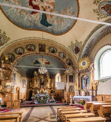 Zdjęcie nr 24: Kościół usytuowany jest w południowo-wschodniej części wsi, otoczony kamiennym murem, otynkowanym, krytym betonową płytą, w który wbudowano współczesne stacje drogi krzyżowej. Na plac kościelny prowadzą trzy bramy. Główna brama znajduje się w murze od strony zachodniej, nakryta jest ostrołukową arkadą. Dwie mniejsze bramy zlokalizowane są od strony południowej oraz od strony wschodniej. Na placu przed kościołem stoi pomnik z 1911 roku wystawiony na pamiątkę księdza Wojciecha Blaszyńskiego i działacza ludowego Stanisława Stojałowskiego. 
Świątynia jest orientowana, murowana, jednonawowa z węższym i krótkim prezbiterium, zamkniętym półkoliście oraz z wieżą na zakończeniu nawy. Do prezbiterium przylega zakrystia od północy i skarbczyk od południa. Od strony południowej dobudowana jest również niska kruchta. Wnętrze trójprzęsłowej nawy rozczłonkowane jest filarami przyściennymi wspierającymi sklepienie żagielkowe. Chór muzyczny trójarkadowy, wsparty na dwóch filarach. W ścianach korpusu nawowego znajdują się głębokie wnęki, w których umieszczone są prostokątne otwory okienne zamknięte półkolistym łukiem, po trzy od strony północnej i południowej; takie same okno znajduje się również w apsydzie od strony południowej oraz na chórze od strony zachodniej. Ponadto w apsydzie na wysokości zwieńczenia ołtarza głównego umieszczone jest małe, okrągłe okno. W kruchcie, skarbczyku i zakrystii okna są prostokątne. Drzwi główne dwuskrzydłowe, drewniane z metalowymi okuciami, zabezpieczone od wewnątrz dwiema żelaznymi zaporami umocowanymi w murze. Podobne drzwi znajdują się od strony południowej z kruchty. 
Fasada kościoła jest płaska, zakończona gzymsem z wysuniętą do przodu trójkondygnacyjną wieżą, nakrytą baniastym hełmem z latarnią. Drugie piętro wieży połączone jest z fasadą prostymi spływami. Przy wejściu na wysokich cokołach ustawione są rzeźby świętych Piotra i Pawła. 
Kościoł nakryty jest dachem dwuspadowym, na skrzyżowaniu nawy z prezbiterium umieszczona jest sygnaturka z hełmem baniastym z latarnią. 
