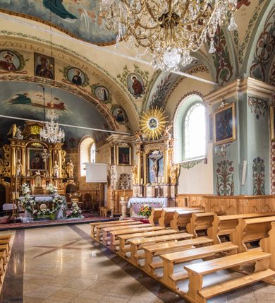 Zdjęcie nr 26: Kościół usytuowany jest w południowo-wschodniej części wsi, otoczony kamiennym murem, otynkowanym, krytym betonową płytą, w który wbudowano współczesne stacje drogi krzyżowej. Na plac kościelny prowadzą trzy bramy. Główna brama znajduje się w murze od strony zachodniej, nakryta jest ostrołukową arkadą. Dwie mniejsze bramy zlokalizowane są od strony południowej oraz od strony wschodniej. Na placu przed kościołem stoi pomnik z 1911 roku wystawiony na pamiątkę księdza Wojciecha Blaszyńskiego i działacza ludowego Stanisława Stojałowskiego. 
Świątynia jest orientowana, murowana, jednonawowa z węższym i krótkim prezbiterium, zamkniętym półkoliście oraz z wieżą na zakończeniu nawy. Do prezbiterium przylega zakrystia od północy i skarbczyk od południa. Od strony południowej dobudowana jest również niska kruchta. Wnętrze trójprzęsłowej nawy rozczłonkowane jest filarami przyściennymi wspierającymi sklepienie żagielkowe. Chór muzyczny trójarkadowy, wsparty na dwóch filarach. W ścianach korpusu nawowego znajdują się głębokie wnęki, w których umieszczone są prostokątne otwory okienne zamknięte półkolistym łukiem, po trzy od strony północnej i południowej; takie same okno znajduje się również w apsydzie od strony południowej oraz na chórze od strony zachodniej. Ponadto w apsydzie na wysokości zwieńczenia ołtarza głównego umieszczone jest małe, okrągłe okno. W kruchcie, skarbczyku i zakrystii okna są prostokątne. Drzwi główne dwuskrzydłowe, drewniane z metalowymi okuciami, zabezpieczone od wewnątrz dwiema żelaznymi zaporami umocowanymi w murze. Podobne drzwi znajdują się od strony południowej z kruchty. 
Fasada kościoła jest płaska, zakończona gzymsem z wysuniętą do przodu trójkondygnacyjną wieżą, nakrytą baniastym hełmem z latarnią. Drugie piętro wieży połączone jest z fasadą prostymi spływami. Przy wejściu na wysokich cokołach ustawione są rzeźby świętych Piotra i Pawła. 
Kościoł nakryty jest dachem dwuspadowym, na skrzyżowaniu nawy z prezbiterium umieszczona jest sygnaturka z hełmem baniastym z latarnią. 
