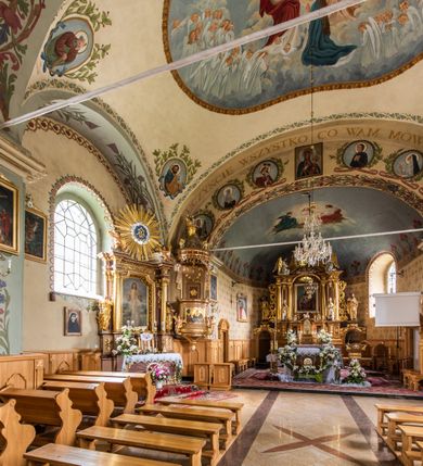 Zdjęcie nr 25: Kościół usytuowany jest w południowo-wschodniej części wsi, otoczony kamiennym murem, otynkowanym, krytym betonową płytą, w który wbudowano współczesne stacje drogi krzyżowej. Na plac kościelny prowadzą trzy bramy. Główna brama znajduje się w murze od strony zachodniej, nakryta jest ostrołukową arkadą. Dwie mniejsze bramy zlokalizowane są od strony południowej oraz od strony wschodniej. Na placu przed kościołem stoi pomnik z 1911 roku wystawiony na pamiątkę księdza Wojciecha Blaszyńskiego i działacza ludowego Stanisława Stojałowskiego. 
Świątynia jest orientowana, murowana, jednonawowa z węższym i krótkim prezbiterium, zamkniętym półkoliście oraz z wieżą na zakończeniu nawy. Do prezbiterium przylega zakrystia od północy i skarbczyk od południa. Od strony południowej dobudowana jest również niska kruchta. Wnętrze trójprzęsłowej nawy rozczłonkowane jest filarami przyściennymi wspierającymi sklepienie żagielkowe. Chór muzyczny trójarkadowy, wsparty na dwóch filarach. W ścianach korpusu nawowego znajdują się głębokie wnęki, w których umieszczone są prostokątne otwory okienne zamknięte półkolistym łukiem, po trzy od strony północnej i południowej; takie same okno znajduje się również w apsydzie od strony południowej oraz na chórze od strony zachodniej. Ponadto w apsydzie na wysokości zwieńczenia ołtarza głównego umieszczone jest małe, okrągłe okno. W kruchcie, skarbczyku i zakrystii okna są prostokątne. Drzwi główne dwuskrzydłowe, drewniane z metalowymi okuciami, zabezpieczone od wewnątrz dwiema żelaznymi zaporami umocowanymi w murze. Podobne drzwi znajdują się od strony południowej z kruchty. 
Fasada kościoła jest płaska, zakończona gzymsem z wysuniętą do przodu trójkondygnacyjną wieżą, nakrytą baniastym hełmem z latarnią. Drugie piętro wieży połączone jest z fasadą prostymi spływami. Przy wejściu na wysokich cokołach ustawione są rzeźby świętych Piotra i Pawła. 
Kościoł nakryty jest dachem dwuspadowym, na skrzyżowaniu nawy z prezbiterium umieszczona jest sygnaturka z hełmem baniastym z latarnią. 
