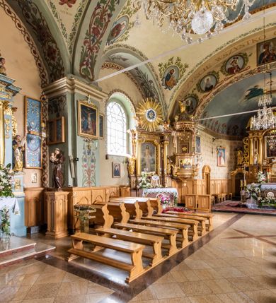 Zdjęcie nr 29: Kościół usytuowany jest w południowo-wschodniej części wsi, otoczony kamiennym murem, otynkowanym, krytym betonową płytą, w który wbudowano współczesne stacje drogi krzyżowej. Na plac kościelny prowadzą trzy bramy. Główna brama znajduje się w murze od strony zachodniej, nakryta jest ostrołukową arkadą. Dwie mniejsze bramy zlokalizowane są od strony południowej oraz od strony wschodniej. Na placu przed kościołem stoi pomnik z 1911 roku wystawiony na pamiątkę księdza Wojciecha Blaszyńskiego i działacza ludowego Stanisława Stojałowskiego. 
Świątynia jest orientowana, murowana, jednonawowa z węższym i krótkim prezbiterium, zamkniętym półkoliście oraz z wieżą na zakończeniu nawy. Do prezbiterium przylega zakrystia od północy i skarbczyk od południa. Od strony południowej dobudowana jest również niska kruchta. Wnętrze trójprzęsłowej nawy rozczłonkowane jest filarami przyściennymi wspierającymi sklepienie żagielkowe. Chór muzyczny trójarkadowy, wsparty na dwóch filarach. W ścianach korpusu nawowego znajdują się głębokie wnęki, w których umieszczone są prostokątne otwory okienne zamknięte półkolistym łukiem, po trzy od strony północnej i południowej; takie same okno znajduje się również w apsydzie od strony południowej oraz na chórze od strony zachodniej. Ponadto w apsydzie na wysokości zwieńczenia ołtarza głównego umieszczone jest małe, okrągłe okno. W kruchcie, skarbczyku i zakrystii okna są prostokątne. Drzwi główne dwuskrzydłowe, drewniane z metalowymi okuciami, zabezpieczone od wewnątrz dwiema żelaznymi zaporami umocowanymi w murze. Podobne drzwi znajdują się od strony południowej z kruchty. 
Fasada kościoła jest płaska, zakończona gzymsem z wysuniętą do przodu trójkondygnacyjną wieżą, nakrytą baniastym hełmem z latarnią. Drugie piętro wieży połączone jest z fasadą prostymi spływami. Przy wejściu na wysokich cokołach ustawione są rzeźby świętych Piotra i Pawła. 
Kościoł nakryty jest dachem dwuspadowym, na skrzyżowaniu nawy z prezbiterium umieszczona jest sygnaturka z hełmem baniastym z latarnią. 
