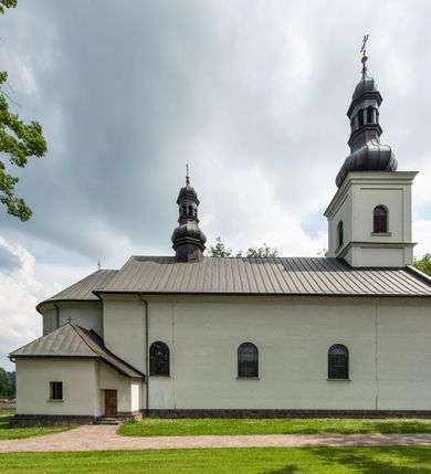 Zdjęcie nr 13: Kościół usytuowany jest w południowo-wschodniej części wsi, otoczony kamiennym murem, otynkowanym, krytym betonową płytą, w który wbudowano współczesne stacje drogi krzyżowej. Na plac kościelny prowadzą trzy bramy. Główna brama znajduje się w murze od strony zachodniej, nakryta jest ostrołukową arkadą. Dwie mniejsze bramy zlokalizowane są od strony południowej oraz od strony wschodniej. Na placu przed kościołem stoi pomnik z 1911 roku wystawiony na pamiątkę księdza Wojciecha Blaszyńskiego i działacza ludowego Stanisława Stojałowskiego. 
Świątynia jest orientowana, murowana, jednonawowa z węższym i krótkim prezbiterium, zamkniętym półkoliście oraz z wieżą na zakończeniu nawy. Do prezbiterium przylega zakrystia od północy i skarbczyk od południa. Od strony południowej dobudowana jest również niska kruchta. Wnętrze trójprzęsłowej nawy rozczłonkowane jest filarami przyściennymi wspierającymi sklepienie żagielkowe. Chór muzyczny trójarkadowy, wsparty na dwóch filarach. W ścianach korpusu nawowego znajdują się głębokie wnęki, w których umieszczone są prostokątne otwory okienne zamknięte półkolistym łukiem, po trzy od strony północnej i południowej; takie same okno znajduje się również w apsydzie od strony południowej oraz na chórze od strony zachodniej. Ponadto w apsydzie na wysokości zwieńczenia ołtarza głównego umieszczone jest małe, okrągłe okno. W kruchcie, skarbczyku i zakrystii okna są prostokątne. Drzwi główne dwuskrzydłowe, drewniane z metalowymi okuciami, zabezpieczone od wewnątrz dwiema żelaznymi zaporami umocowanymi w murze. Podobne drzwi znajdują się od strony południowej z kruchty. 
Fasada kościoła jest płaska, zakończona gzymsem z wysuniętą do przodu trójkondygnacyjną wieżą, nakrytą baniastym hełmem z latarnią. Drugie piętro wieży połączone jest z fasadą prostymi spływami. Przy wejściu na wysokich cokołach ustawione są rzeźby świętych Piotra i Pawła. 
Kościoł nakryty jest dachem dwuspadowym, na skrzyżowaniu nawy z prezbiterium umieszczona jest sygnaturka z hełmem baniastym z latarnią. 
