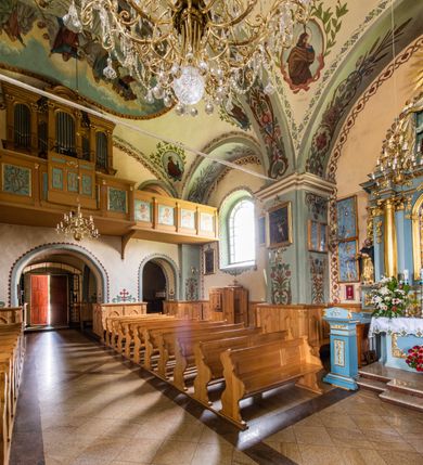 Zdjęcie nr 31: Kościół usytuowany jest w południowo-wschodniej części wsi, otoczony kamiennym murem, otynkowanym, krytym betonową płytą, w który wbudowano współczesne stacje drogi krzyżowej. Na plac kościelny prowadzą trzy bramy. Główna brama znajduje się w murze od strony zachodniej, nakryta jest ostrołukową arkadą. Dwie mniejsze bramy zlokalizowane są od strony południowej oraz od strony wschodniej. Na placu przed kościołem stoi pomnik z 1911 roku wystawiony na pamiątkę księdza Wojciecha Blaszyńskiego i działacza ludowego Stanisława Stojałowskiego. 
Świątynia jest orientowana, murowana, jednonawowa z węższym i krótkim prezbiterium, zamkniętym półkoliście oraz z wieżą na zakończeniu nawy. Do prezbiterium przylega zakrystia od północy i skarbczyk od południa. Od strony południowej dobudowana jest również niska kruchta. Wnętrze trójprzęsłowej nawy rozczłonkowane jest filarami przyściennymi wspierającymi sklepienie żagielkowe. Chór muzyczny trójarkadowy, wsparty na dwóch filarach. W ścianach korpusu nawowego znajdują się głębokie wnęki, w których umieszczone są prostokątne otwory okienne zamknięte półkolistym łukiem, po trzy od strony północnej i południowej; takie same okno znajduje się również w apsydzie od strony południowej oraz na chórze od strony zachodniej. Ponadto w apsydzie na wysokości zwieńczenia ołtarza głównego umieszczone jest małe, okrągłe okno. W kruchcie, skarbczyku i zakrystii okna są prostokątne. Drzwi główne dwuskrzydłowe, drewniane z metalowymi okuciami, zabezpieczone od wewnątrz dwiema żelaznymi zaporami umocowanymi w murze. Podobne drzwi znajdują się od strony południowej z kruchty. 
Fasada kościoła jest płaska, zakończona gzymsem z wysuniętą do przodu trójkondygnacyjną wieżą, nakrytą baniastym hełmem z latarnią. Drugie piętro wieży połączone jest z fasadą prostymi spływami. Przy wejściu na wysokich cokołach ustawione są rzeźby świętych Piotra i Pawła. 
Kościoł nakryty jest dachem dwuspadowym, na skrzyżowaniu nawy z prezbiterium umieszczona jest sygnaturka z hełmem baniastym z latarnią. 
