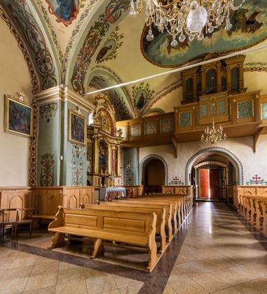 Zdjęcie nr 33: Kościół usytuowany jest w południowo-wschodniej części wsi, otoczony kamiennym murem, otynkowanym, krytym betonową płytą, w który wbudowano współczesne stacje drogi krzyżowej. Na plac kościelny prowadzą trzy bramy. Główna brama znajduje się w murze od strony zachodniej, nakryta jest ostrołukową arkadą. Dwie mniejsze bramy zlokalizowane są od strony południowej oraz od strony wschodniej. Na placu przed kościołem stoi pomnik z 1911 roku wystawiony na pamiątkę księdza Wojciecha Blaszyńskiego i działacza ludowego Stanisława Stojałowskiego. 
Świątynia jest orientowana, murowana, jednonawowa z węższym i krótkim prezbiterium, zamkniętym półkoliście oraz z wieżą na zakończeniu nawy. Do prezbiterium przylega zakrystia od północy i skarbczyk od południa. Od strony południowej dobudowana jest również niska kruchta. Wnętrze trójprzęsłowej nawy rozczłonkowane jest filarami przyściennymi wspierającymi sklepienie żagielkowe. Chór muzyczny trójarkadowy, wsparty na dwóch filarach. W ścianach korpusu nawowego znajdują się głębokie wnęki, w których umieszczone są prostokątne otwory okienne zamknięte półkolistym łukiem, po trzy od strony północnej i południowej; takie same okno znajduje się również w apsydzie od strony południowej oraz na chórze od strony zachodniej. Ponadto w apsydzie na wysokości zwieńczenia ołtarza głównego umieszczone jest małe, okrągłe okno. W kruchcie, skarbczyku i zakrystii okna są prostokątne. Drzwi główne dwuskrzydłowe, drewniane z metalowymi okuciami, zabezpieczone od wewnątrz dwiema żelaznymi zaporami umocowanymi w murze. Podobne drzwi znajdują się od strony południowej z kruchty. 
Fasada kościoła jest płaska, zakończona gzymsem z wysuniętą do przodu trójkondygnacyjną wieżą, nakrytą baniastym hełmem z latarnią. Drugie piętro wieży połączone jest z fasadą prostymi spływami. Przy wejściu na wysokich cokołach ustawione są rzeźby świętych Piotra i Pawła. 
Kościoł nakryty jest dachem dwuspadowym, na skrzyżowaniu nawy z prezbiterium umieszczona jest sygnaturka z hełmem baniastym z latarnią. 
