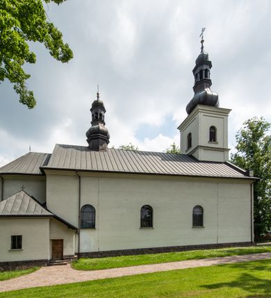 Zdjęcie nr 16: Kościół usytuowany jest w południowo-wschodniej części wsi, otoczony kamiennym murem, otynkowanym, krytym betonową płytą, w który wbudowano współczesne stacje drogi krzyżowej. Na plac kościelny prowadzą trzy bramy. Główna brama znajduje się w murze od strony zachodniej, nakryta jest ostrołukową arkadą. Dwie mniejsze bramy zlokalizowane są od strony południowej oraz od strony wschodniej. Na placu przed kościołem stoi pomnik z 1911 roku wystawiony na pamiątkę księdza Wojciecha Blaszyńskiego i działacza ludowego Stanisława Stojałowskiego. 
Świątynia jest orientowana, murowana, jednonawowa z węższym i krótkim prezbiterium, zamkniętym półkoliście oraz z wieżą na zakończeniu nawy. Do prezbiterium przylega zakrystia od północy i skarbczyk od południa. Od strony południowej dobudowana jest również niska kruchta. Wnętrze trójprzęsłowej nawy rozczłonkowane jest filarami przyściennymi wspierającymi sklepienie żagielkowe. Chór muzyczny trójarkadowy, wsparty na dwóch filarach. W ścianach korpusu nawowego znajdują się głębokie wnęki, w których umieszczone są prostokątne otwory okienne zamknięte półkolistym łukiem, po trzy od strony północnej i południowej; takie same okno znajduje się również w apsydzie od strony południowej oraz na chórze od strony zachodniej. Ponadto w apsydzie na wysokości zwieńczenia ołtarza głównego umieszczone jest małe, okrągłe okno. W kruchcie, skarbczyku i zakrystii okna są prostokątne. Drzwi główne dwuskrzydłowe, drewniane z metalowymi okuciami, zabezpieczone od wewnątrz dwiema żelaznymi zaporami umocowanymi w murze. Podobne drzwi znajdują się od strony południowej z kruchty. 
Fasada kościoła jest płaska, zakończona gzymsem z wysuniętą do przodu trójkondygnacyjną wieżą, nakrytą baniastym hełmem z latarnią. Drugie piętro wieży połączone jest z fasadą prostymi spływami. Przy wejściu na wysokich cokołach ustawione są rzeźby świętych Piotra i Pawła. 
Kościoł nakryty jest dachem dwuspadowym, na skrzyżowaniu nawy z prezbiterium umieszczona jest sygnaturka z hełmem baniastym z latarnią. 
