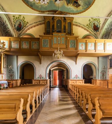 Zdjęcie nr 34: Kościół usytuowany jest w południowo-wschodniej części wsi, otoczony kamiennym murem, otynkowanym, krytym betonową płytą, w który wbudowano współczesne stacje drogi krzyżowej. Na plac kościelny prowadzą trzy bramy. Główna brama znajduje się w murze od strony zachodniej, nakryta jest ostrołukową arkadą. Dwie mniejsze bramy zlokalizowane są od strony południowej oraz od strony wschodniej. Na placu przed kościołem stoi pomnik z 1911 roku wystawiony na pamiątkę księdza Wojciecha Blaszyńskiego i działacza ludowego Stanisława Stojałowskiego. 
Świątynia jest orientowana, murowana, jednonawowa z węższym i krótkim prezbiterium, zamkniętym półkoliście oraz z wieżą na zakończeniu nawy. Do prezbiterium przylega zakrystia od północy i skarbczyk od południa. Od strony południowej dobudowana jest również niska kruchta. Wnętrze trójprzęsłowej nawy rozczłonkowane jest filarami przyściennymi wspierającymi sklepienie żagielkowe. Chór muzyczny trójarkadowy, wsparty na dwóch filarach. W ścianach korpusu nawowego znajdują się głębokie wnęki, w których umieszczone są prostokątne otwory okienne zamknięte półkolistym łukiem, po trzy od strony północnej i południowej; takie same okno znajduje się również w apsydzie od strony południowej oraz na chórze od strony zachodniej. Ponadto w apsydzie na wysokości zwieńczenia ołtarza głównego umieszczone jest małe, okrągłe okno. W kruchcie, skarbczyku i zakrystii okna są prostokątne. Drzwi główne dwuskrzydłowe, drewniane z metalowymi okuciami, zabezpieczone od wewnątrz dwiema żelaznymi zaporami umocowanymi w murze. Podobne drzwi znajdują się od strony południowej z kruchty. 
Fasada kościoła jest płaska, zakończona gzymsem z wysuniętą do przodu trójkondygnacyjną wieżą, nakrytą baniastym hełmem z latarnią. Drugie piętro wieży połączone jest z fasadą prostymi spływami. Przy wejściu na wysokich cokołach ustawione są rzeźby świętych Piotra i Pawła. 
Kościoł nakryty jest dachem dwuspadowym, na skrzyżowaniu nawy z prezbiterium umieszczona jest sygnaturka z hełmem baniastym z latarnią. 
