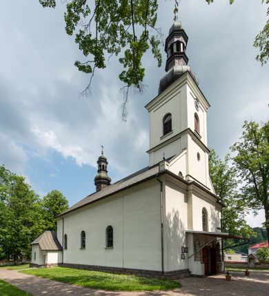 Zdjęcie nr 15: Kościół usytuowany jest w południowo-wschodniej części wsi, otoczony kamiennym murem, otynkowanym, krytym betonową płytą, w który wbudowano współczesne stacje drogi krzyżowej. Na plac kościelny prowadzą trzy bramy. Główna brama znajduje się w murze od strony zachodniej, nakryta jest ostrołukową arkadą. Dwie mniejsze bramy zlokalizowane są od strony południowej oraz od strony wschodniej. Na placu przed kościołem stoi pomnik z 1911 roku wystawiony na pamiątkę księdza Wojciecha Blaszyńskiego i działacza ludowego Stanisława Stojałowskiego. 
Świątynia jest orientowana, murowana, jednonawowa z węższym i krótkim prezbiterium, zamkniętym półkoliście oraz z wieżą na zakończeniu nawy. Do prezbiterium przylega zakrystia od północy i skarbczyk od południa. Od strony południowej dobudowana jest również niska kruchta. Wnętrze trójprzęsłowej nawy rozczłonkowane jest filarami przyściennymi wspierającymi sklepienie żagielkowe. Chór muzyczny trójarkadowy, wsparty na dwóch filarach. W ścianach korpusu nawowego znajdują się głębokie wnęki, w których umieszczone są prostokątne otwory okienne zamknięte półkolistym łukiem, po trzy od strony północnej i południowej; takie same okno znajduje się również w apsydzie od strony południowej oraz na chórze od strony zachodniej. Ponadto w apsydzie na wysokości zwieńczenia ołtarza głównego umieszczone jest małe, okrągłe okno. W kruchcie, skarbczyku i zakrystii okna są prostokątne. Drzwi główne dwuskrzydłowe, drewniane z metalowymi okuciami, zabezpieczone od wewnątrz dwiema żelaznymi zaporami umocowanymi w murze. Podobne drzwi znajdują się od strony południowej z kruchty. 
Fasada kościoła jest płaska, zakończona gzymsem z wysuniętą do przodu trójkondygnacyjną wieżą, nakrytą baniastym hełmem z latarnią. Drugie piętro wieży połączone jest z fasadą prostymi spływami. Przy wejściu na wysokich cokołach ustawione są rzeźby świętych Piotra i Pawła. 
Kościoł nakryty jest dachem dwuspadowym, na skrzyżowaniu nawy z prezbiterium umieszczona jest sygnaturka z hełmem baniastym z latarnią. 

