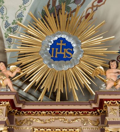 Zdjęcie nr 48: Nastawa ołtarzowa architektoniczna, jednoosiowa i jednokondygnacyjna. Ołtarz prostopadłościenny, drewniany, z drewnianą mensą i z marmurowym portatylem. Antepedium w formie prostokąta ujętego profilowaną ramą. W polu płycina o narożach zdobionych płaskorzeźbioną dekoracją roślinną złożoną z liści stylizowanego akantu oraz umieszczonym w polu veraikonem na tle obłoków i w promienistej glorii. Tabernakulum w kształcie prostopadłościennej skrzynki na niskim cokole, flankowane dwiema kolumnami ustawionymi pod kątem, zamknięte od góry prostym belkowaniem, pośrodku nadwieszonym. Na drzwiczkach płycina w kształcie stojącego prostokąta, zamkniętego łukiem półkolistym, ujęta profilowaną ramą, w polu której umieszczony jest płaskorzeźbiony kielich z hostią.

Retabulum ustawione na podwójnym cokole, flankowane parą zdwojonych pilastrów i ustawionych przed nimi dwiema kolumnami. Podpory dźwigają wyłamane na osi belkowanie z ustawionymi na bokach rzeźbami aniołków z arma Christi (włóczniami). Kolumny wspierają się na bazach, trzony mają gładkie i złocone, a kapitele kompozytowe. Po bokach ołtarza na niskich cokołach ustawione są rzeźby aniołów z narzędziami męki Pańskiej w dłoniach: kolumną biczowania po lewej stronie i włócznią po prawej stronie. W polu, w głębokiej wnęce ujętej szeroką, profilowaną ramą umieszczony jest późnobarokowy krucyfiks na tle malowanej Golgoty i postaci Matki Boskiej, św. Jana Ewangelisty i św. Weroniki. Wnęka od wewnątrz zdobiona jest płaskorzeźbioną winną latoroślą. Cokół dekorowany jest prostokątnymi po bokach płycinami, wypełnionymi stylizowanym akantem. Belkowanie zdobione wicią liści akantu, imposty rozetkami. W zwieńczeniu monumentalna promienista gloria złożona z wiązek promieni o nierównej długości z monogramem Chrystusa na niebieskim tle w polu, otoczonym wieńcem z obłoków. Struktura polichromowana w kolorze jasnego, delikatnego fioletu, płyciny, filary i detale w odcieniu delikatnie ciemniejszym; ornamentyka i profilowania złocone i srebrzone.


