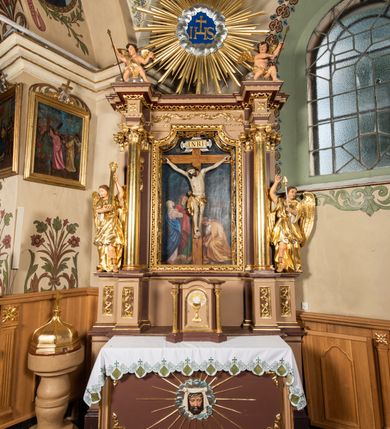 Zdjęcie nr 3: Nastawa ołtarzowa architektoniczna, jednoosiowa i jednokondygnacyjna. Ołtarz prostopadłościenny, drewniany, z drewnianą mensą i z marmurowym portatylem. Antepedium w formie prostokąta ujętego profilowaną ramą. W polu płycina o narożach zdobionych płaskorzeźbioną dekoracją roślinną złożoną z liści stylizowanego akantu oraz umieszczonym w polu veraikonem na tle obłoków i w promienistej glorii. Tabernakulum w kształcie prostopadłościennej skrzynki na niskim cokole, flankowane dwiema kolumnami ustawionymi pod kątem, zamknięte od góry prostym belkowaniem, pośrodku nadwieszonym. Na drzwiczkach płycina w kształcie stojącego prostokąta, zamkniętego łukiem półkolistym, ujęta profilowaną ramą, w polu której umieszczony jest płaskorzeźbiony kielich z hostią.

Retabulum ustawione na podwójnym cokole, flankowane parą zdwojonych pilastrów i ustawionych przed nimi dwiema kolumnami. Podpory dźwigają wyłamane na osi belkowanie z ustawionymi na bokach rzeźbami aniołków z arma Christi (włóczniami). Kolumny wspierają się na bazach, trzony mają gładkie i złocone, a kapitele kompozytowe. Po bokach ołtarza na niskich cokołach ustawione są rzeźby aniołów z narzędziami męki Pańskiej w dłoniach: kolumną biczowania po lewej stronie i włócznią po prawej stronie. W polu, w głębokiej wnęce ujętej szeroką, profilowaną ramą umieszczony jest późnobarokowy krucyfiks na tle malowanej Golgoty i postaci Matki Boskiej, św. Jana Ewangelisty i św. Weroniki. Wnęka od wewnątrz zdobiona jest płaskorzeźbioną winną latoroślą. Cokół dekorowany jest prostokątnymi po bokach płycinami, wypełnionymi stylizowanym akantem. Belkowanie zdobione wicią liści akantu, imposty rozetkami. W zwieńczeniu monumentalna promienista gloria złożona z wiązek promieni o nierównej długości z monogramem Chrystusa na niebieskim tle w polu, otoczonym wieńcem z obłoków. Struktura polichromowana w kolorze jasnego, delikatnego fioletu, płyciny, filary i detale w odcieniu delikatnie ciemniejszym; ornamentyka i profilowania złocone i srebrzone.


