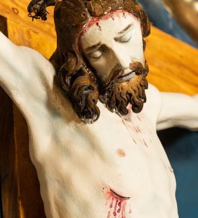 Zdjęcie nr 11: Krucyfiks w typie Cristo morte; postać  Chrystusa umarłego przybitego do krzyża trzema gwoździami. Sylwetka w delikatnym zwisie, o silnie zaznaczonej anatomii ciała, z podkreśloną linią żeber i zaznaczonymi śladami męki. Perizonium w formie tkaniny przewiązanej na sznurze na prawym boku, sięgające kolan i odsłaniające prawe udo. Twarz Jezusa podłużna opadająca na prawe ramię, okolona zarostem; oczy zamknięte, nos podłużny, usta delikatnie otwarte; włosy długie spływające na prawe ramię, na głowie zielona korona cierniowa. Krzyż drewniany, prosty. Nad głową Chrystusa zawieszona tabliczka w kształcie kartusza zdobionego ornamentem okuciowym z napisem „I.N.R.I.” Partie ciała polichromowane naturalistycznie, perizonium złocone.




