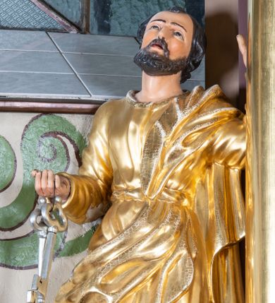 Zdjęcie nr 1: Rzeźba św. Piotra, drążona, ustawiona na niskim prostopadłościennym cokole w kolorze jasnobrązowym. Święty ustawiony frontalnie z głową uniesioną delikatnie do góry. W prawej ręce, ugiętej w łokciu trzyma dwa srebrne klucze, lewą rękę opiera o kolumnę. Twarz pociągła o wyrazistych rysach z krótkim ciemnobrązowym zarostem. Święty ubrany jest w długą, przewiązaną w pasie suknię i przerzucony przez lewe ramię złocony płaszcz. Na stopach ma złocone sandały. Polichromia w odsłoniętych partiach ciała naturalistyczna. 