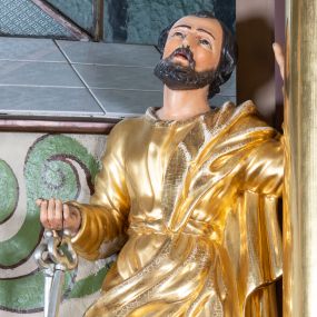 Zdjęcie nr 1: Rzeźba św. Piotra, drążona, ustawiona na niskim prostopadłościennym cokole w kolorze jasnobrązowym. Święty ustawiony frontalnie z głową uniesioną delikatnie do góry. W prawej ręce, ugiętej w łokciu trzyma dwa srebrne klucze, lewą rękę opiera o kolumnę. Twarz pociągła o wyrazistych rysach z krótkim ciemnobrązowym zarostem. Święty ubrany jest w długą, przewiązaną w pasie suknię i przerzucony przez lewe ramię złocony płaszcz. Na stopach ma złocone sandały. Polichromia w odsłoniętych partiach ciała naturalistyczna. 