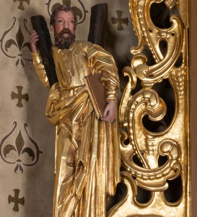 Zdjęcie nr 1: Rzeźba św. Andrzeja ścięta z tyłu i drążona, ustawiona na półkolistym i zielonym cokole. Święty ukazany jest frontalnie w delikatnym kontrapoście. W lewej dłoni trzyma, przytulając do biodra, księgę, a prawą obejmuje i opiera się o belkę krzyża w kształcie litery „X” stojącego za nim. Twarz pociągła z dużym nosem i szeroko rozstawionymi oczami, okolona długą i gęstą brodą oraz krótkimi i kręconymi włosami. Święty ubrany jest w suknię przewiązaną w talii z wąskimi rękawami i kołnierzykiem, zapiętą u góry na cztery guziki, a także płaszcz przewiązany przez lewe ramię i opadający na prawą stronę wzdłuż bioder; na stopach ma sandały. Tkaniny obficie drapowane w proste fałdy, płaszcz z wywiniętym na prawym boku brzegiem. Polichromia naturalistyczna w odsłoniętych partiach ciała, szaty złocone.